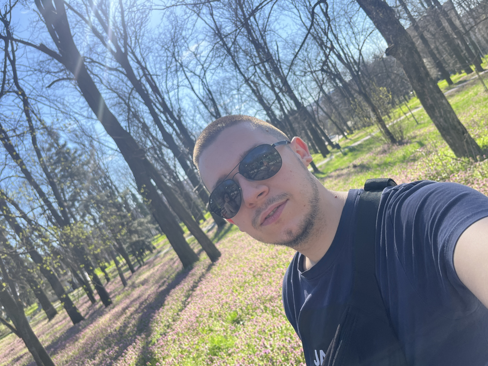 A medium shot depicts a man with a short haircut and sunglasses in a park setting. The man is wearing a dark blue t-shirt. The park features trees and ground cover with small pink flowers. The location is likely in the city of Razgrad, Bulgaria, based on the reflection in the sunglasses. The trees are largely bare, suggesting early spring. The sky is blue with some light flares from the sun.