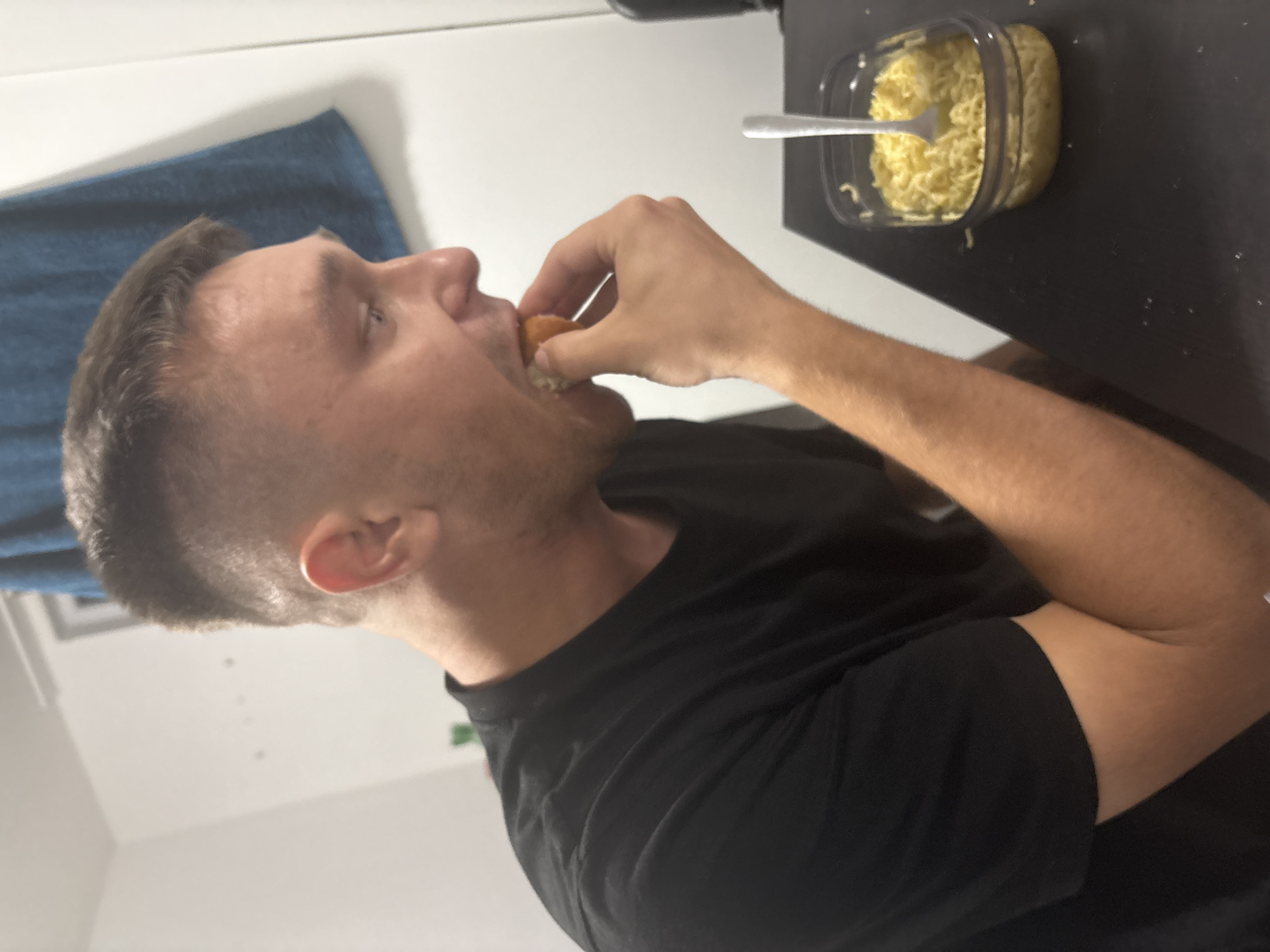 A man is seated at a black table, eating instant ramen noodles from a clear plastic container. He is wearing a black t-shirt. A silver fork rests in the ramen. A blue towel hangs in the background against a white wall. The man is in the process of putting a piece of food into his mouth with his right hand.