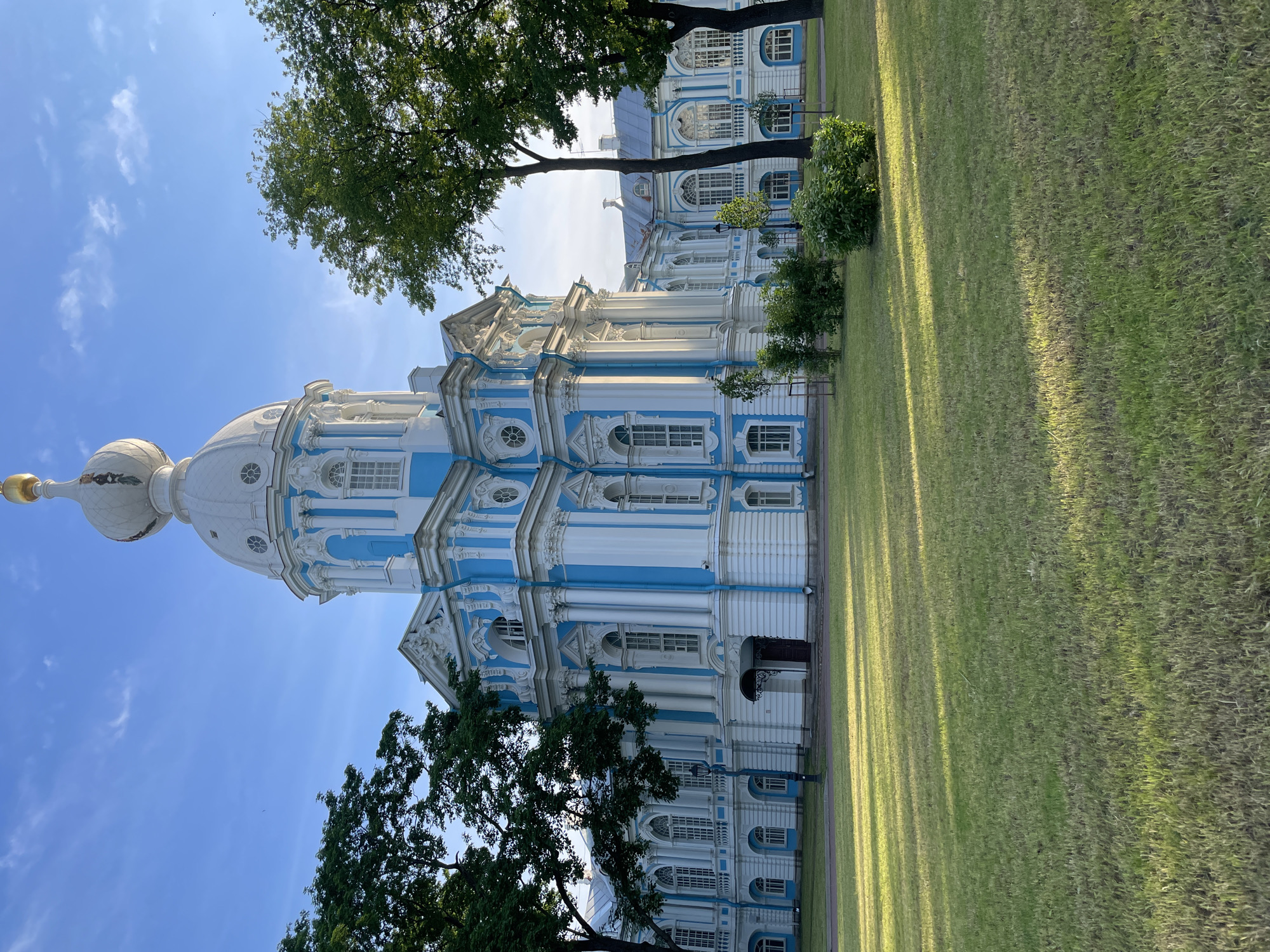 This image presents a wide exterior view of the Smolny Cathedral, located in St. Petersburg, Russia, under a clear, bright blue sky with sparse, wispy white clouds. The grand, ornate building dominates the upper and central portions of the frame.
The cathedral is constructed in a distinct Baroque style, characterized by its striking white facade intricately accented with light blue pilasters, window frames, cornices, and decorative elements. The central feature is a large, multi-tiered white dome topped by a smaller, white onion-shaped cupola which, in turn, is capped by a prominent golden onion dome. Visible on the white cupola, just below the gold, are decorative patterns or figures. Below the main dome, several small, round windows are symmetrically arranged. The building features numerous large, tall windows with white frames, many of which are arched, and some have multiple panes. White, often fluted, columns and pilasters contribute to the architectural detailing on various sections of the facade. A dark entrance door, sheltered by a small, dark canopy, is visible on the central part of the building. To the right, extensions of the cathedral complex are visible, maintaining the same blue and white color scheme and architectural style, with grey or light blue/grey pitched roofs.
In the foreground, a large, well-maintained expanse of green grass extends across the lower half of the image. Long, distinct shadows from unseen objects to the left, as well as from the visible trees and perhaps the building itself, stretch diagonally across the grass, suggesting morning or late afternoon sunlight. A dark-colored path or road runs along the edge of the grass closer to the building.
Lush, green deciduous trees frame the building on both the left and right sides, partially obscuring the lower portions of the structure. Smaller green bushes and saplings are also present in the grassy area closer to the building. A dark lamppost stands near the path on the left side of the scene. No individuals are visible in the image. The scene is brightly illuminated by natural sunlight, casting vibrant colors on the building and grounds.