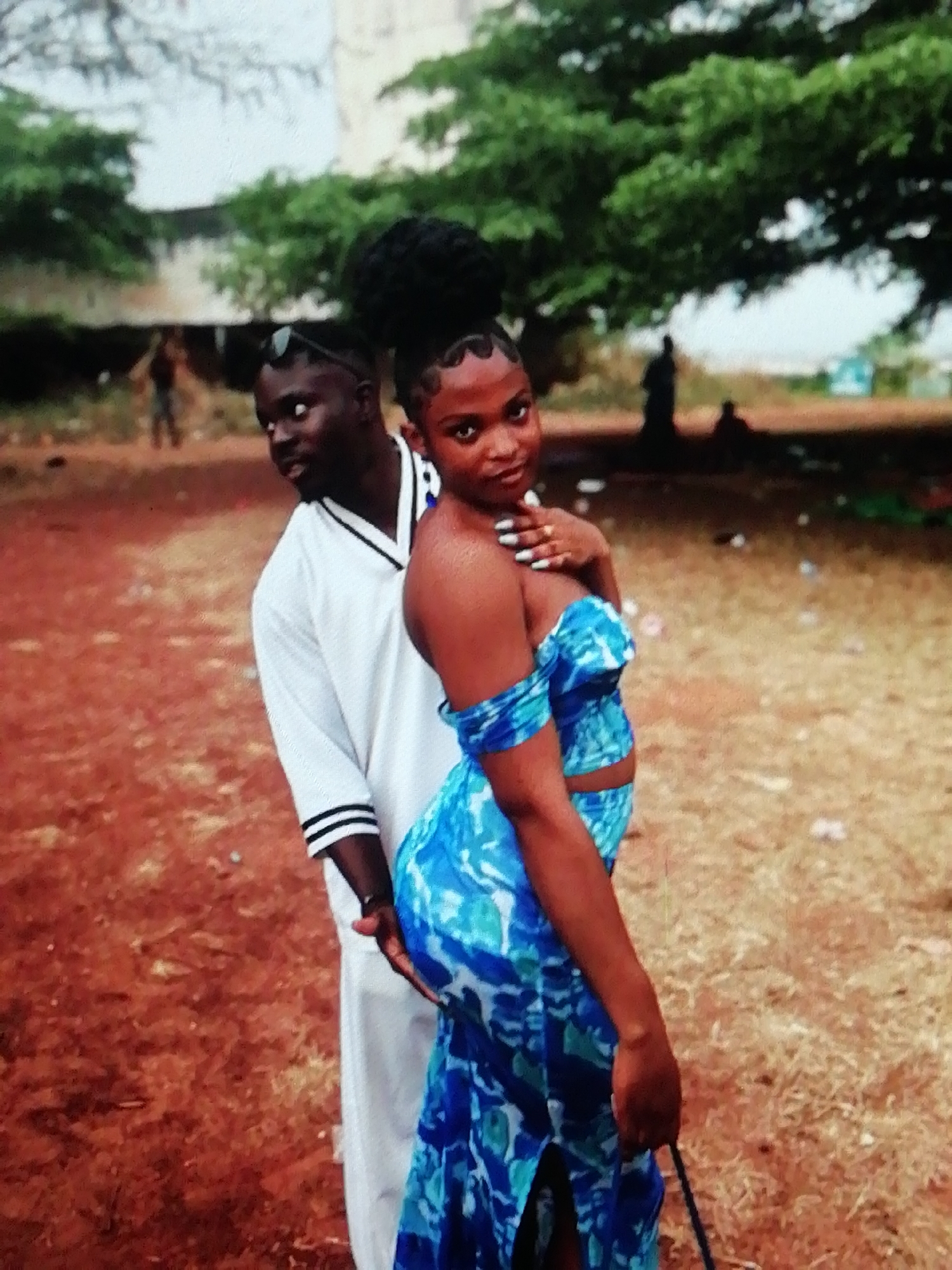 The image shows a dark-skinned woman in a blue, patterned two-piece dress posing in front of a dark-skinned man wearing a white V-neck shirt with black stripes. The man stands behind the woman, with his left hand appearing to rest on her hip. The woman has dark hair styled up, and her left hand is near her face. The scene is set outdoors on what appears to be red soil. In the background, there are trees and indistinct figures. While it is difficult to precisely pinpoint the exact location, the photo is likely taken in Enugu, Nigeria, given the prompt context and the environmental and physical characteristics of the subjects.