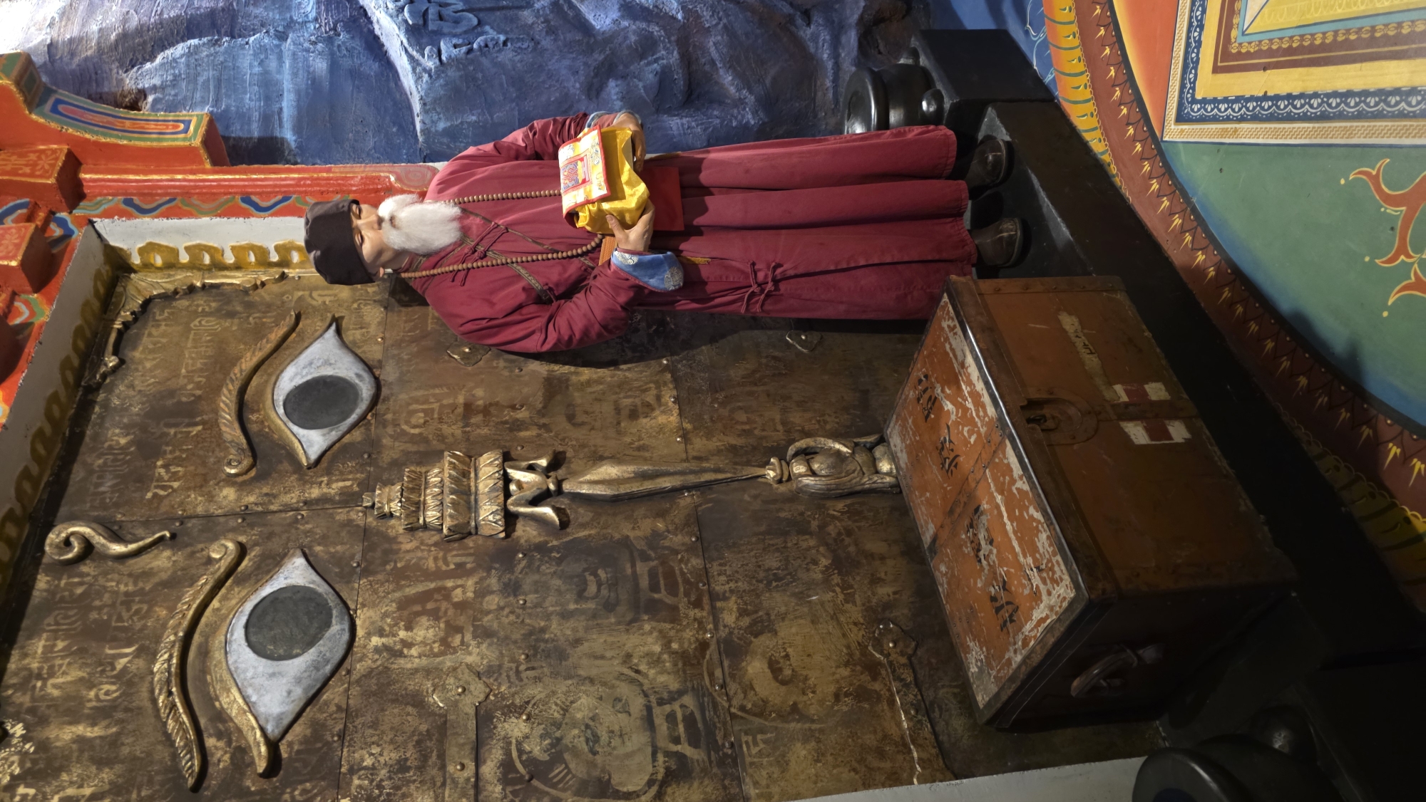 A sideways view shows a wax statue of a man in a burgundy robe with a white beard and black hat. He is holding a gold object and wearing prayer beads. The man is lying on top of a large gold door with two eye emblems. To the right of the door is an aged orange trunk with painted text. The door and statue are inside a temple on Vasilievsky Island in Saint Petersburg, Russia.