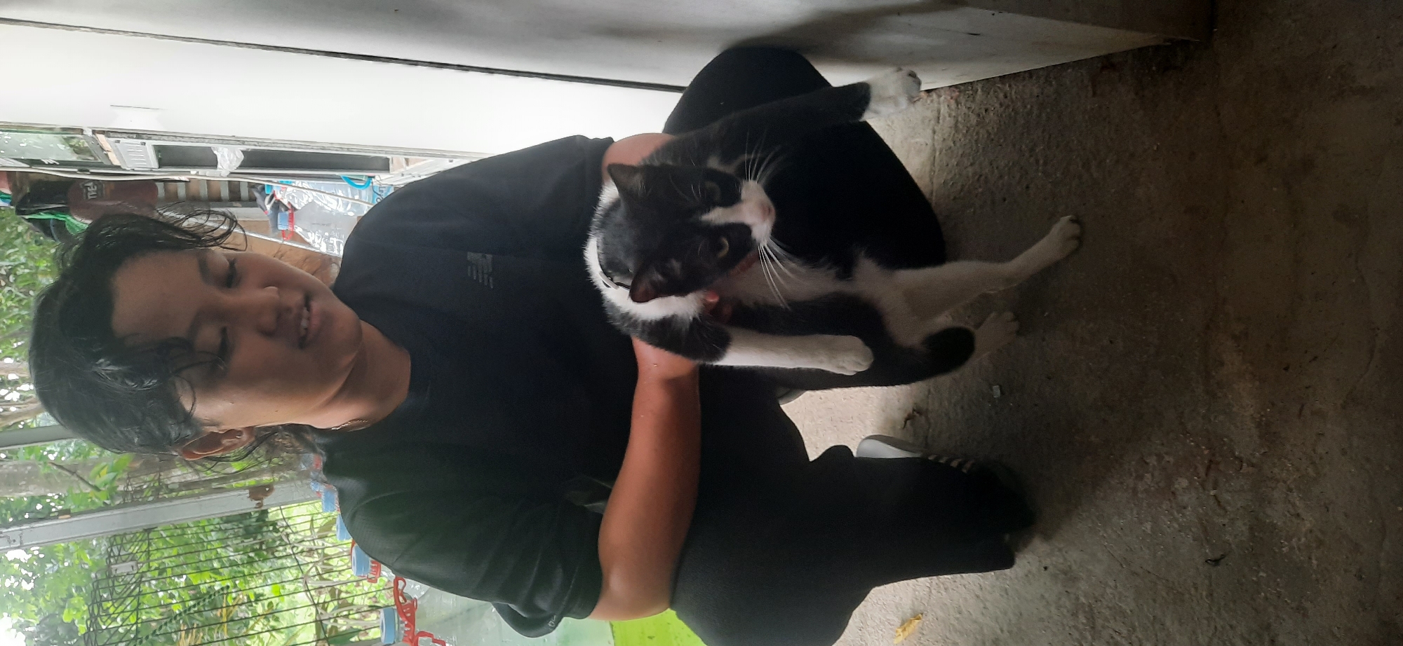 A person is crouched down on a concrete floor, holding a black and white cat. The person is wearing a black t-shirt and black pants. The cat is being held upside down, with its front paws hanging down. The background of the image shows a fence with greenery behind it, as well as a white wall and some bottles. The lighting suggests it is daytime. The scene appears to be outdoors or in an open-air structure. The location is identified as Cisauk, Indonesia.