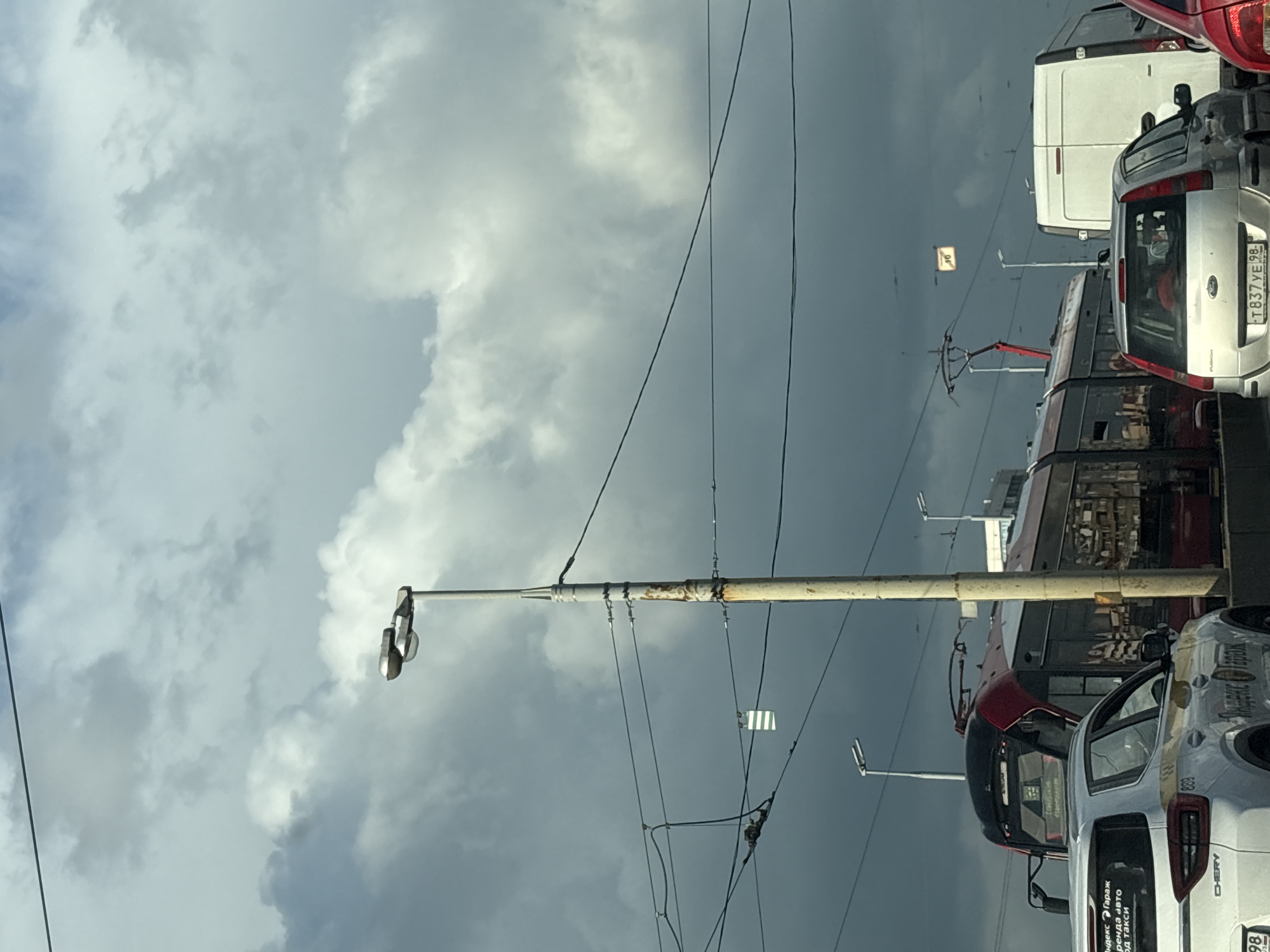 The image depicts a street scene in Saint Petersburg, Russia, with a tram and several vehicles on a cloudy day. A tall, weathered lamppost with two lamps stands prominently in the foreground, partially obscured by electrical wires. To the left, the front of a red and white tram is visible, with some text on its rear window. A white car with "Yandex.Taxi" branding is parked in front of the tram. Further to the right, a white van and the back of a silver car are visible. Several streetlights and overhead electrical wires for the tram system are present throughout the scene. A small, yellow sign with black markings is suspended from the wires in the upper right portion of the image. The sky is filled with dramatic, dark clouds.