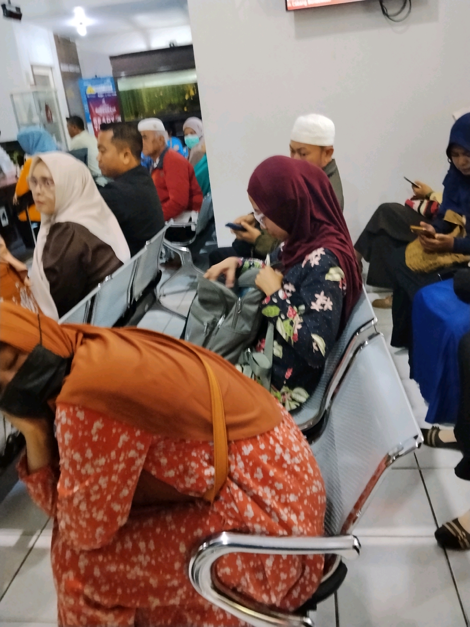 The image shows a waiting area, likely at a public facility or clinic, with several people seated on a row of chairs. The chairs are metallic and have a modern, functional design. The people in the image are dressed in various attire, with several individuals wearing hijabs, suggesting a context where Islamic dress is common. One person in the foreground is wearing an orange top and a red and white floral patterned dress. Another person is wearing a maroon hijab and a floral print top. A man with a white skullcap is visible in the middle ground, and further back, an elderly man in a red jacket is seated. The environment appears to be indoors, with fluorescent lighting and plain walls. A poster or banner is partially visible in the background. The overall impression is one of a typical waiting room scene. The image does not provide specific visual cues to pinpoint the location within Bondowoso, Indonesia, beyond it being an indoor public space.