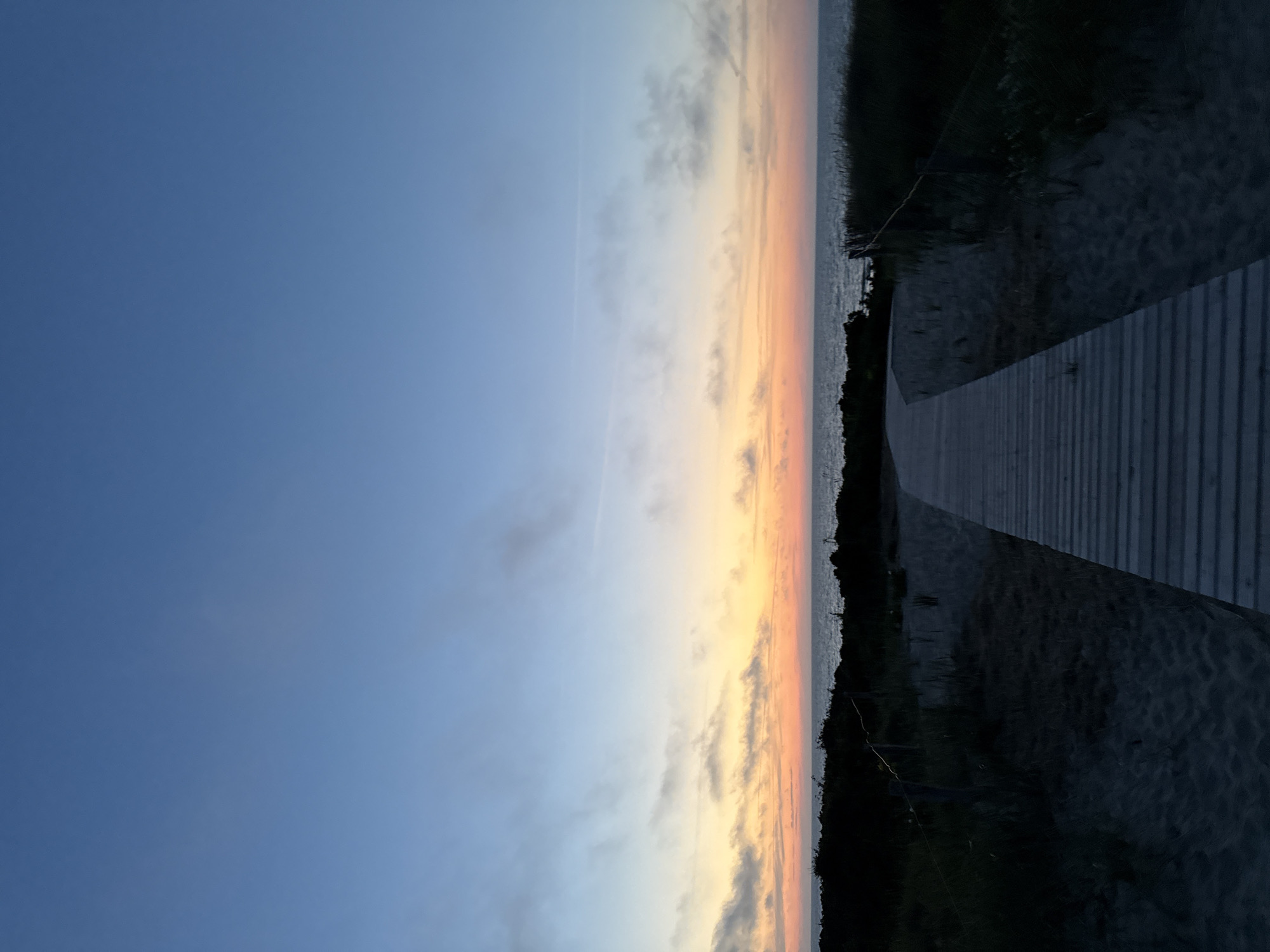 The image depicts a serene scene at sunset on the island of Langeoog, Germany. A wooden boardwalk, made of weathered planks, curves gently through sandy terrain towards the ocean. The boardwalk is framed by dune grass and low-lying vegetation, casting dark shadows in the fading light. In the distance, the calm sea meets the horizon, where the sky transitions from a deep blue at the zenith to soft hues of orange and pink near the water, indicating the sun has recently set. Wispy clouds are scattered across the sky, catching the last remnants of sunlight. The overall atmosphere is tranquil and natural, characteristic of a coastal landscape at dusk.