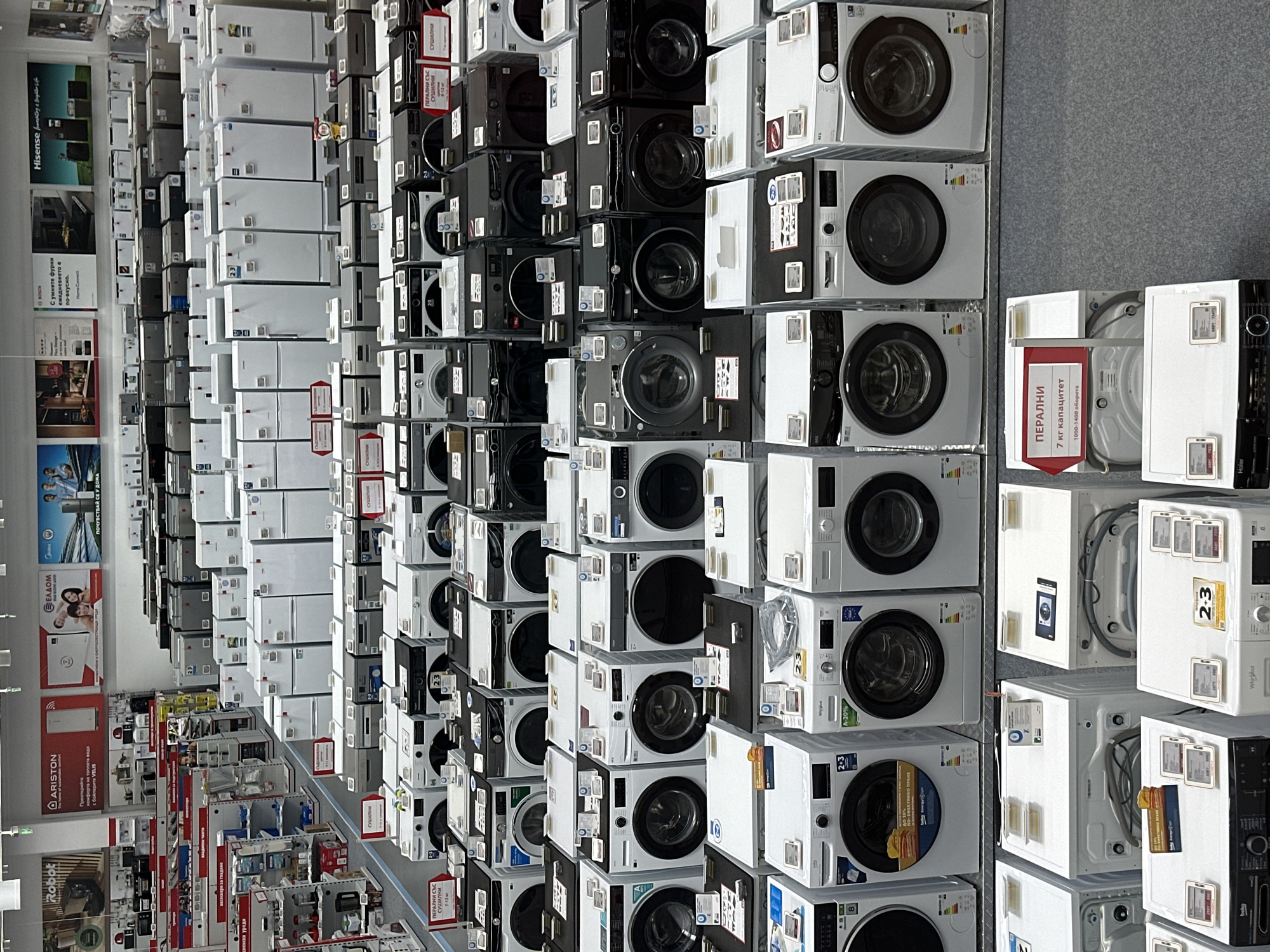 An extensive array of home appliances fills an indoor retail space, likely an electronics store or showroom in Lovech, Bulgaria. The image is rotated clockwise, showing numerous washing machines and refrigerators stacked high.
On the left, rows of white refrigerators, mostly identical in design, are neatly arranged from floor to ceiling. Adjacent to these, a broad selection of front-loading washing machines dominates the center and right side of the frame. These appliances vary in color, including white, black, and silver, and are also stacked in multiple tiers, creating a dense display. Many washing machines feature round doors, control panels, and energy efficiency labels. Some are integrated with dryers.
While no customers or staff are visible, the setup clearly indicates a sales environment. Promotional posters and information tags are attached to walls and individual appliances. The floor appears to be covered with gray carpeting or tile.
Notable text in Cyrillic is prominent, confirming the Bulgarian location. Signs read "ПЕРАЛНИ СЪС Сушилни" (Washing Machines with Dryers), with specifications like "7-12 кг" (7-12 kg). Another tag specifies "ПЕРАЛНИ" (Washing Machines), "7 кг капацитет" (7 kg capacity), and "1000-1400 оборота" (1000-1400 rpm). Brand names like "Hisense," "BOSCH," "ARISTON," "Whirlpool," "Haier," "ELDOM," and "Robot" are visible. A Hisense slogan reads "Smart living & Brighten life," and a Bosch sign says "С умните фурни ежедневието е по-вкусно." (With smart ovens, everyday life is tastier.) Energy ratings like "A-20%" and product numbers such as "2-3" are also frequently seen.