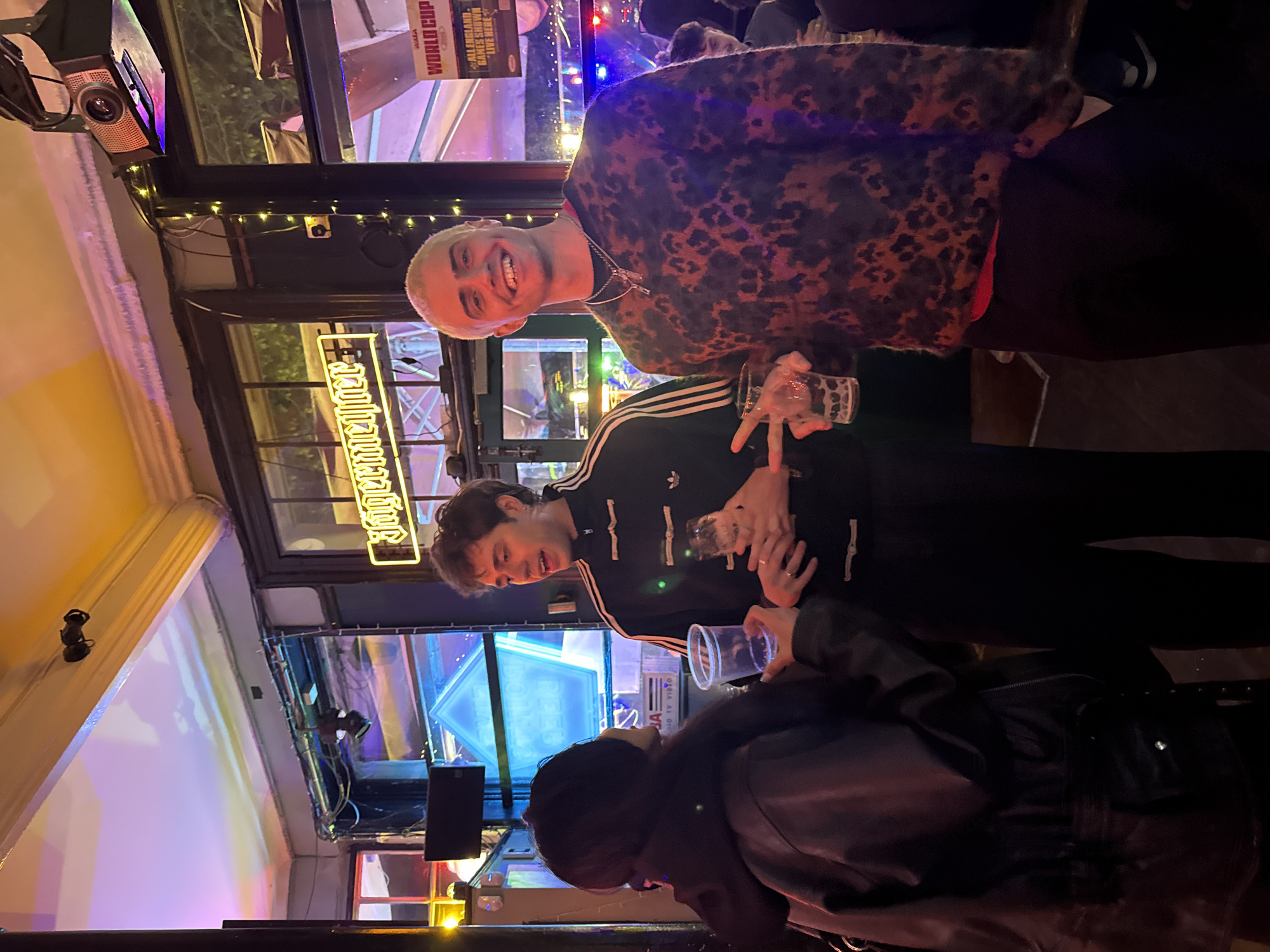 Three individuals are captured inside what appears to be a bar or pub, indicated by the "Jägermeister" sign above the entrance.
One person is wearing a leopard-print sweater and a chain necklace, holding a glass, and smiling. Another is wearing a black Adidas tracksuit. There's a third person with dark hair and a black leather jacket facing away from the camera. All three individuals are interacting socially, suggesting that they may be together.
The environment is brightly lit with ambient lighting, including string lights and colorful light projections. A screen and some advertising materials are visible in the background.
Based on the "World Cup" sign displayed, the location could be a sports bar in the city of London, United Kingdom.
