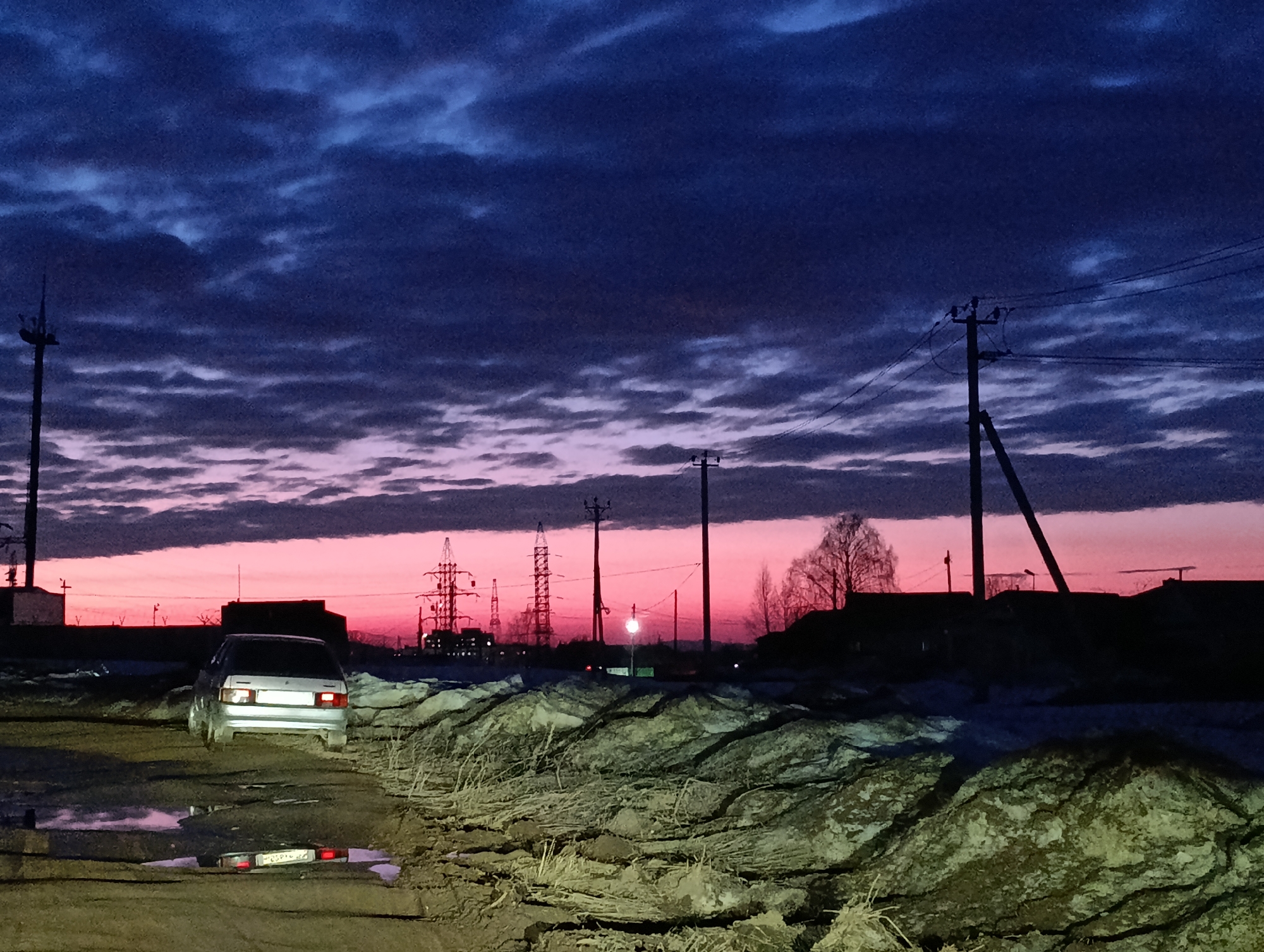 The scene depicts an outdoor environment in Chaikovsky, Perm Krai, Russia, at dawn or dusk. The sky overhead is dark blue with stratified clouds, transitioning to a bright pink and red horizon line. In the foreground, an unpaved, muddy track is visible, bordered by large, irregular mounds of dark soil mixed with dry vegetation and patches of snow. Several puddles are present on the track; one in the immediate foreground reflects the illuminated Russian license plate "P038PH 59 RUS" and the red taillights of a vehicle. A light-colored, four-door hatchback vehicle, consistent with a VAZ-2109 model, is positioned on the track, facing away from the viewer, with its taillights actively illuminated. The ground directly behind and around the vehicle is illuminated by a direct light source. The background features a silhouette of industrial and utility infrastructure against the bright horizon, including a tall mast on the far left, multiple metal lattice power pylons, and numerous utility poles carrying overhead lines. A single bright street lamp or similar light source is visible in the mid-distance. To the right, the silhouettes of bare trees and low-rise buildings are discernible, indicating a mixed industrial-residential area.