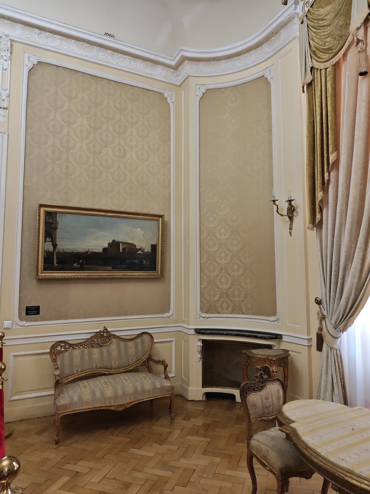 The image depicts an ornate interior room, likely part of a historical building or museum, with elements suggesting an 18th or 19th-century aesthetic. The walls are adorned with a light beige damask wallpaper featuring a repeating circular pattern. Elaborate white stucco molding frames the wall panels and the ceiling.
Dominating the scene is a landscape painting in a gilded frame, hung on the left wall. The painting depicts an architectural scene with classical elements, a body of water, and figures, under a cloudy sky. Below the painting, a small informational plaque is visible.
In the foreground, a light-colored, striped upholstered sofa with ornate gilded wood trim is positioned against the wall. To the right, a matching armchair sits next to a small, carved gilded table. Further to the right, a table with a similar striped top and gilded edge is partially visible. Heavy velvet curtains, in shades of gold and beige, are drawn to the right, framing a window or doorway. A wall-mounted brass candelabra with two light fixtures is present on the right wall. A decorative metal grille is visible below the right-hand wall panel. The floor is made of polished parquet flooring arranged in a herringbone pattern. The scene is well-lit, suggesting ambient interior lighting.
This room is located in the Potocki Palace in Lviv, Ukraine.