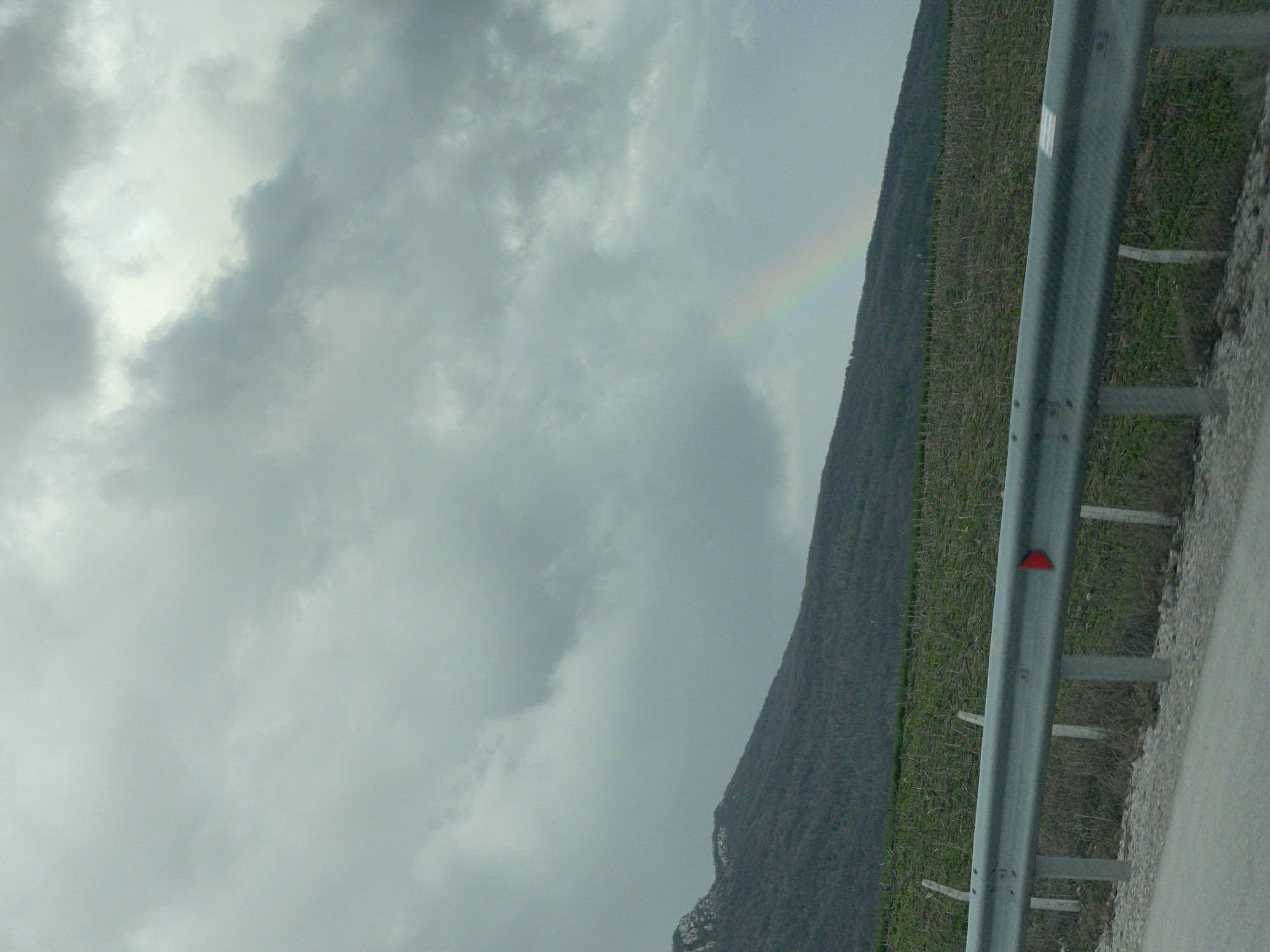 The image depicts an overcast sky with gray clouds and a faint rainbow appearing near a hillside. The hillside is covered in vegetation. In the foreground is a gray metal guardrail, situated beside a road in Buhovtsi, Bulgaria. A red reflector is visible on the guardrail.