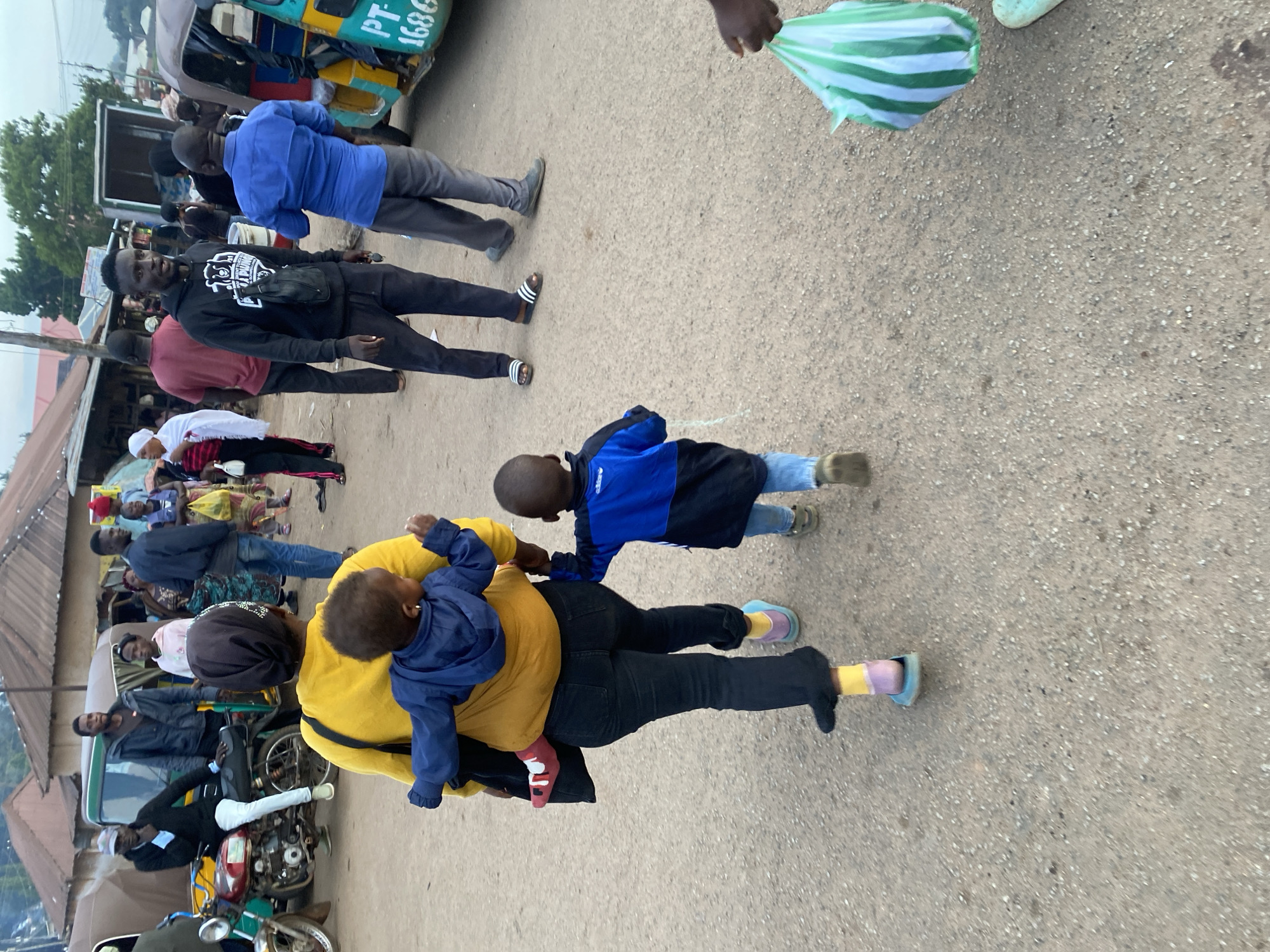 In the city of Bukuru, Nigeria, a woman wearing a yellow top and black pants is seen carrying a baby on her back in a mustard-colored sling. She is walking on a paved road with a young child in a blue and black jacket walking beside her. Several other people are visible in the background, including a man in a black hoodie and dark pants, and others standing near a green and yellow three-wheeled vehicle. The environment appears to be a marketplace or busy street with several buildings and shops lining the road.