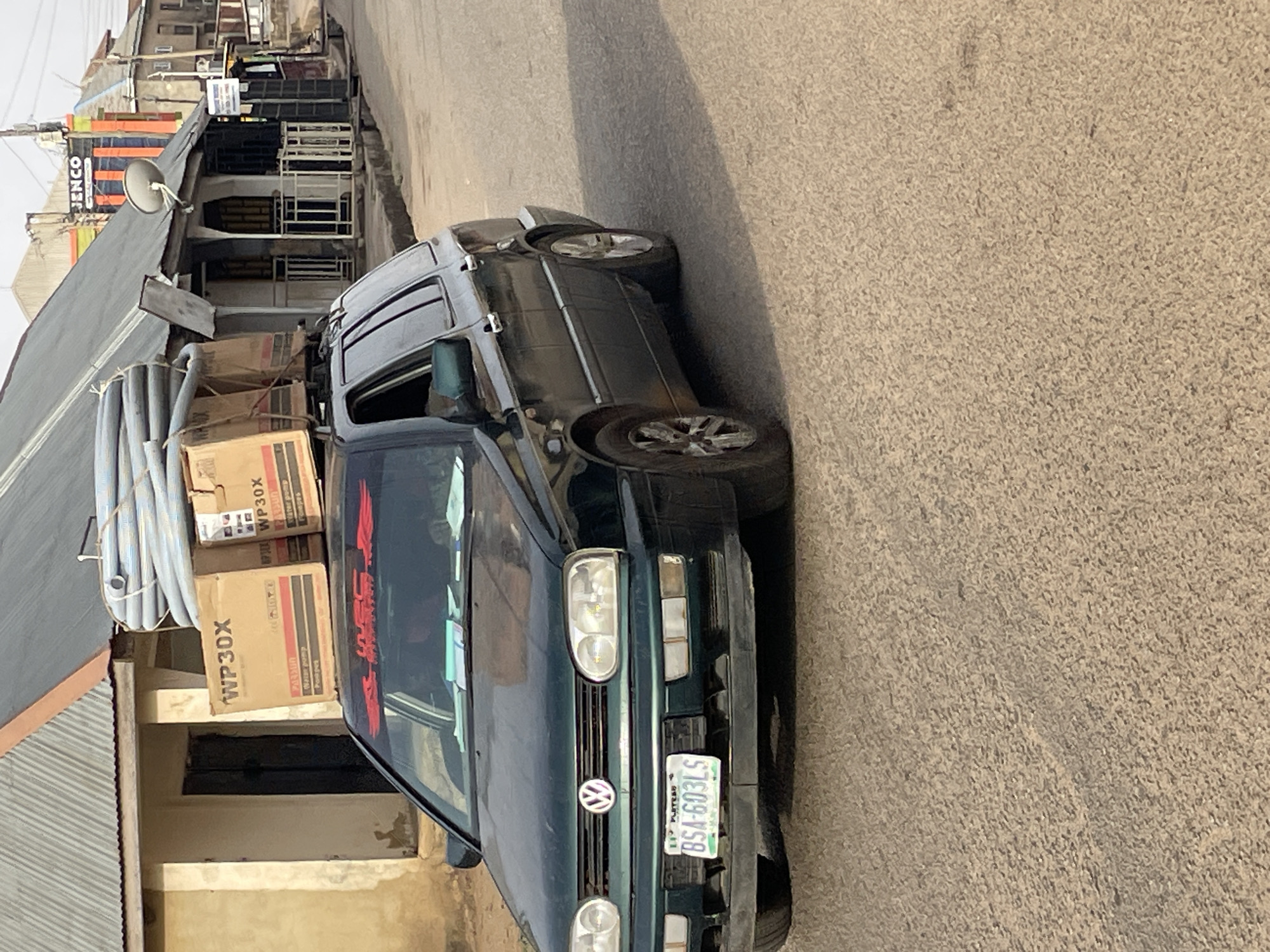 A green Volkswagen car is parked on a paved road in what appears to be a residential or commercial area of Jos, Nigeria. The car has a Nigerian license plate visible: BSA-603LS. The car is heavily loaded with cardboard boxes marked "WP30X" and bundles of what appears to be gray plastic tubing, strapped to its roof. Buildings with corrugated metal roofs and painted facades are visible in the background. A sign that reads "JENCO" is seen in the background. The lighting suggests it is daytime.