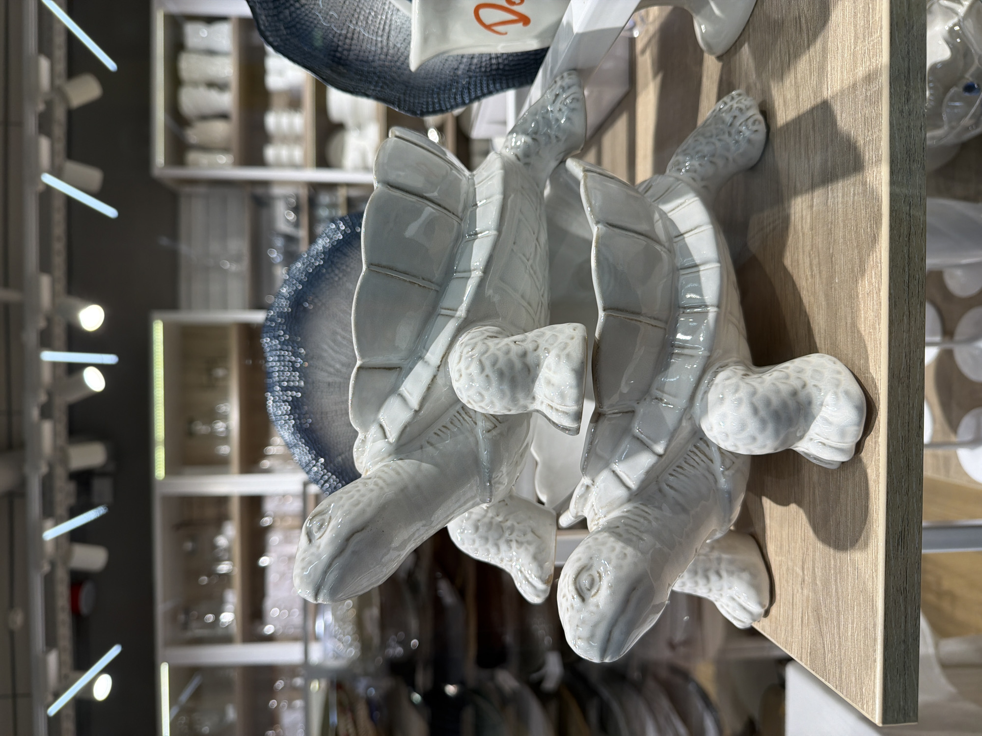 Two white ceramic turtle figurines are stacked on top of each other, placed on a light-colored wooden surface. The figurines are detailed with textured shells and heads. The background shows shelves filled with various items, seemingly in a store or display setting. Due to the lack of additional information, the specific location cannot be determined with certainty. The city of Murmansk, Russia cannot be confirmed as the location.