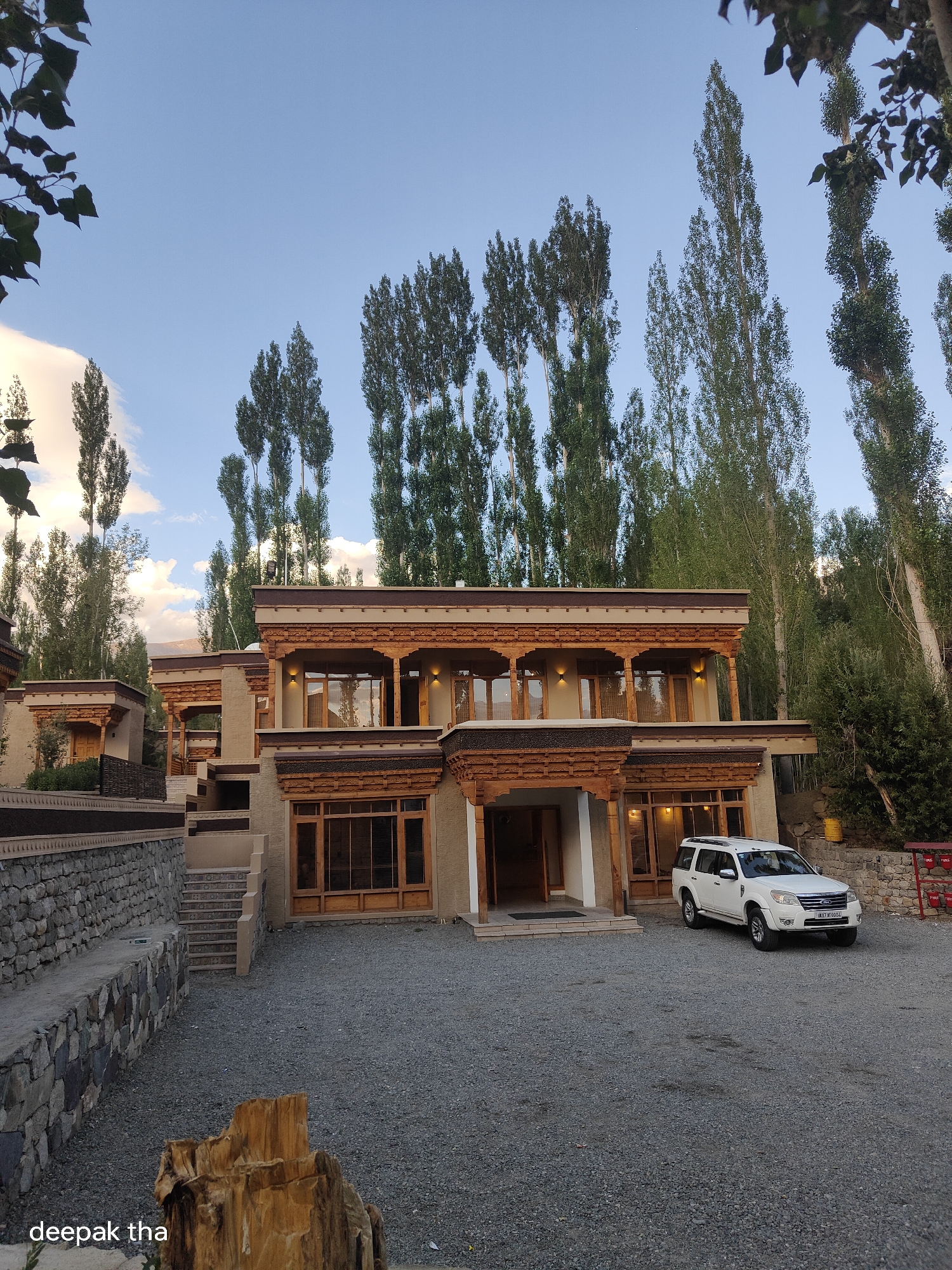 A white SUV is parked on a gravel driveway in front of a multi-story building with traditional architecture. The building features wooden accents, ornate carvings, and large windows. Tall, slender trees line the background, reaching towards a sky with scattered clouds. A stone wall runs along the left side of the property. The water mark "deepak tha" is present in the lower-left corner of the image. The scene appears to be set in Leh, India, given the architectural style and the vegetation.