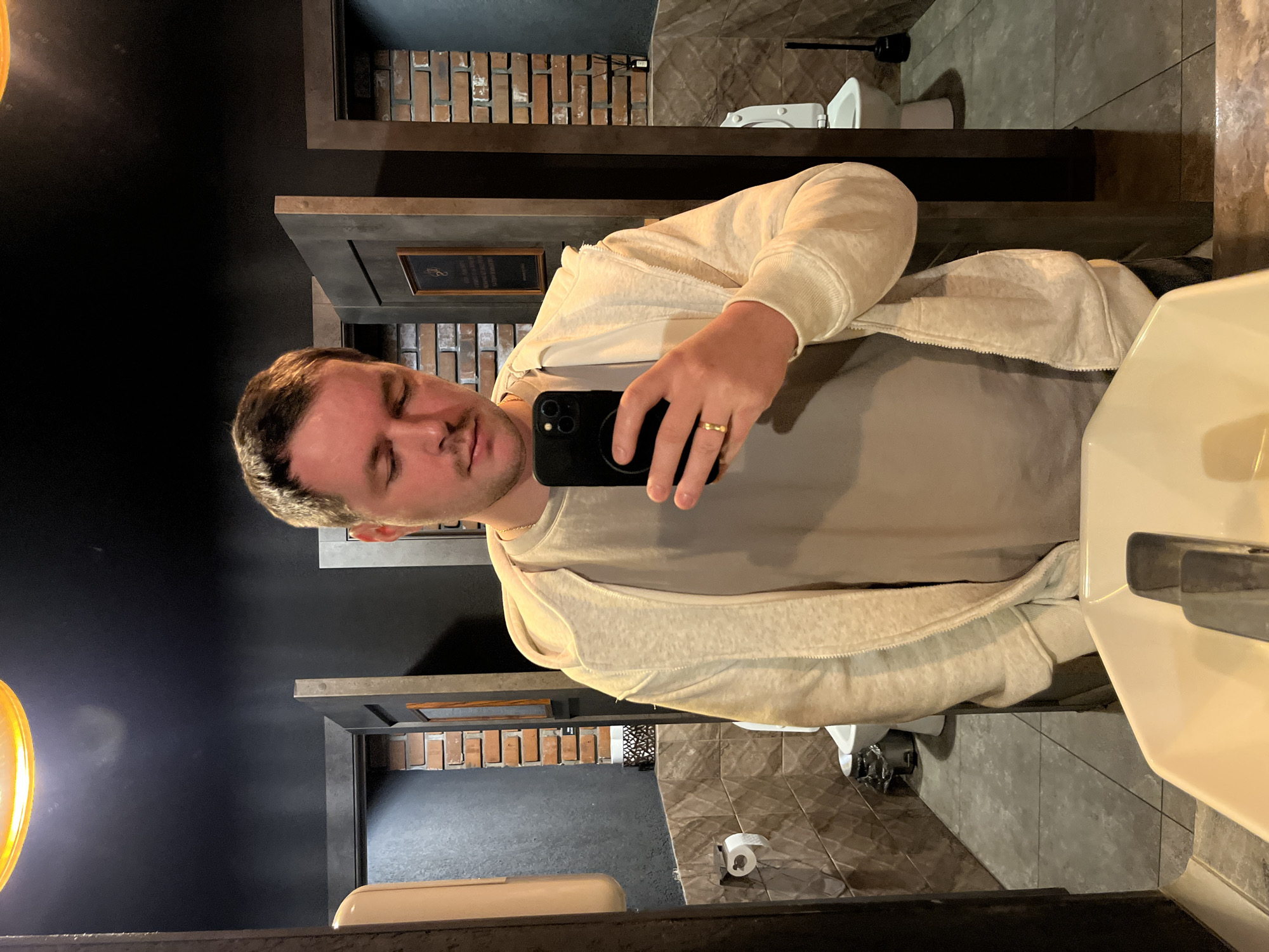 This mirror selfie captures a man in a modern-style restroom, with the image rotated 90 degrees counter-clockwise. The man has short brown hair and appears to have his eyes closed, possibly in a blink or a relaxed moment. He is wearing a light gray t-shirt layered under a cream or light beige hooded zippered sweatshirt. On his left hand, he wears a gold-colored ring and holds a black smartphone with a triple camera setup, which he is using to take the picture.
The setting is a contemporary bathroom, characterized by dark walls, some sections featuring a textured brick-like pattern and others being smooth. The floor is covered in large, possibly gray, textured tiles. In the reflection behind the man, a white ceramic toilet and a roll of toilet paper on a dispenser are visible. A white sink is partially seen on the right side of the frame. The walls incorporate dark shelves or recessed frames, one of which holds a small, framed plaque with illegible text. A warm, yellow-toned light fixture is visible on the far left, casting a glow. The overall aesthetic suggests a well-maintained commercial or public restroom located in Akulovo (Gp Kubinka), Russia.