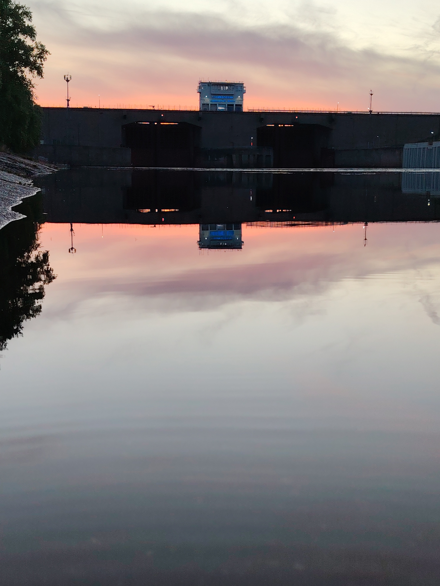 The image depicts the Votkinsk hydroelectric power plant dam in Chaikovsky, Russia, at sunset. The dam, a large concrete structure with several archways, spans across the frame, with a building atop. Reflected in the calm water is the orange and pink sky, the dam itself, and other elements of the landscape. A tree is partially visible on the left. The foreground consists mainly of the water's surface.