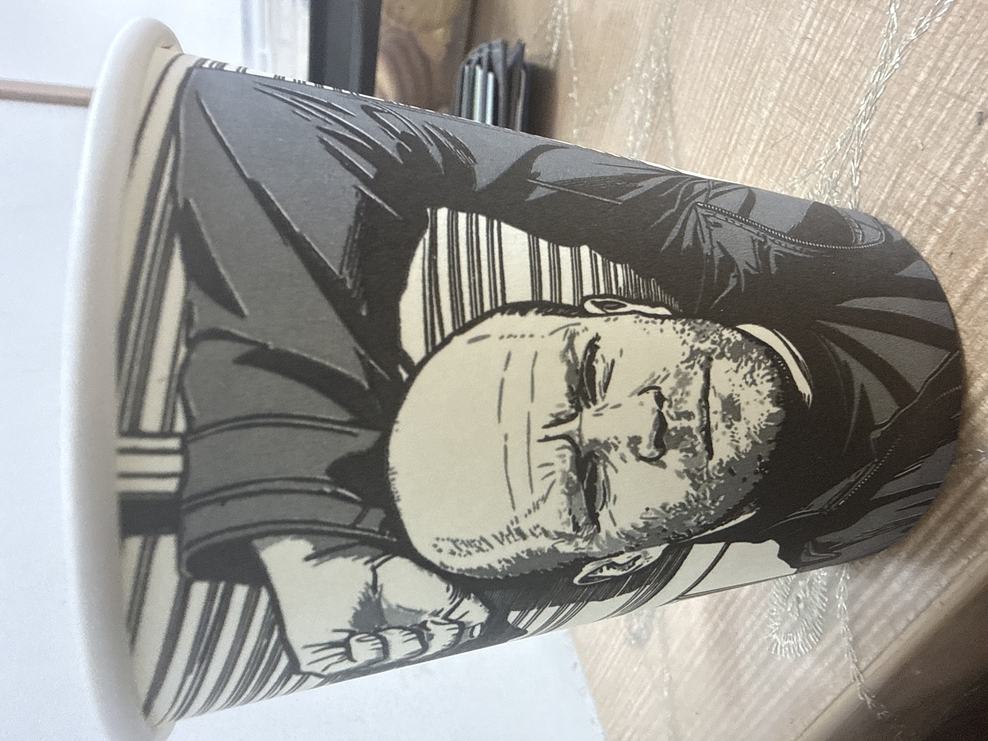 A close-up, eye-level, medium shot shows a disposable coffee cup with a black and white comic-style illustration of a bald man with a beard, wearing a dark shirt. His head is tilted slightly forward, and his eyebrows are furrowed, suggesting a serious or determined expression. A large, drawn fist is raised behind and above his head, partially obscuring the background. The cup is placed on a light brown wooden surface, possibly a table, which has a subtle wood grain pattern. The background is blurred, but appears to be an indoor setting with a light-colored wall. There is no indication of the city of Beslan, Russia, or any specific location within it in this image.
