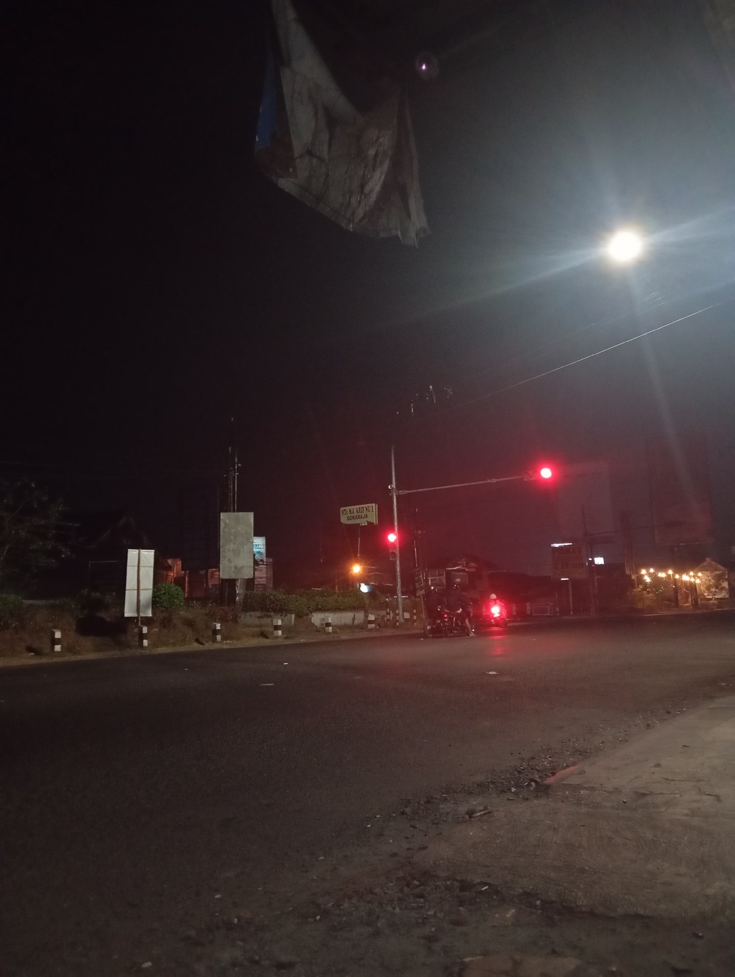 This image captures a nighttime scene at a street intersection in Sokaraja Wetan, Indonesia. The foreground is dominated by a dark asphalt road. A traffic light hangs overhead, displaying a red signal, and another red light is visible lower down on a pole to the left. Two motorcycles with riders are stopped at the intersection, illuminated by the red traffic light. In the background, there are several signs, including one that reads "MTs. MA'ARIF NU 1 SOKARAJA." Buildings with lit windows and decorative string lights are visible on the right side of the intersection, suggesting a commercial or residential area. The sky is dark, with a bright light source, possibly a street lamp or a distant bulb, shining from the upper right. A piece of weathered fabric is suspended from above on the left side of the frame. The overall atmosphere is one of quiet waiting at a traffic signal in a dimly lit urban environment.