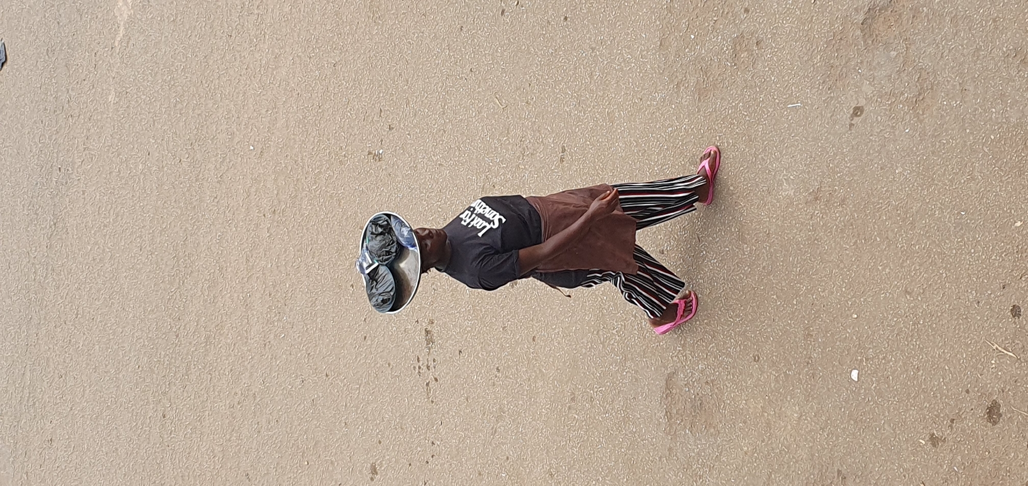 A woman walks across a paved surface in Bukuru, Nigeria, carrying a metal tray balanced on her head. The tray contains several dark plastic bags and some plastic bottles. She is wearing a black t-shirt with white lettering that reads "Last For Somethin," a brown apron, and black and white striped pants. Her feet are adorned with pink flip-flops. The ground is a textured, light brown asphalt.