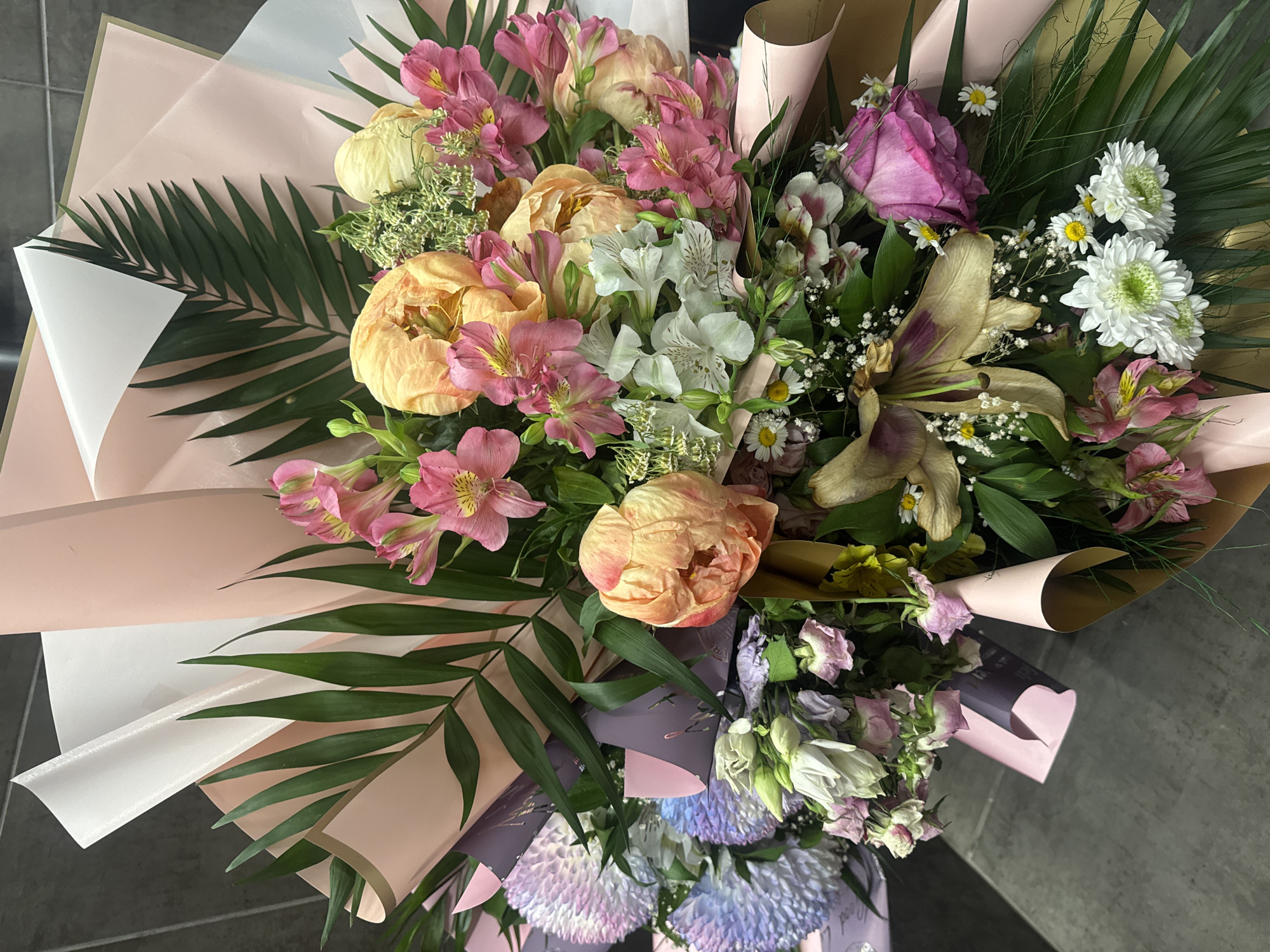 A high-angle, close-up shot showcases a bouquet of flowers. The arrangement features a mix of colors, including pink, peach, white, and purple blossoms, interspersed with green foliage. The flowers are wrapped in layers of pink, white, and kraft-colored paper. There are no people visible in the image. The setting appears to be indoors, possibly in Varna, Bulgaria, though specific landmarks aren't visible to confirm the location. The background is a dark gray, textured surface, possibly a tiled floor or countertop.