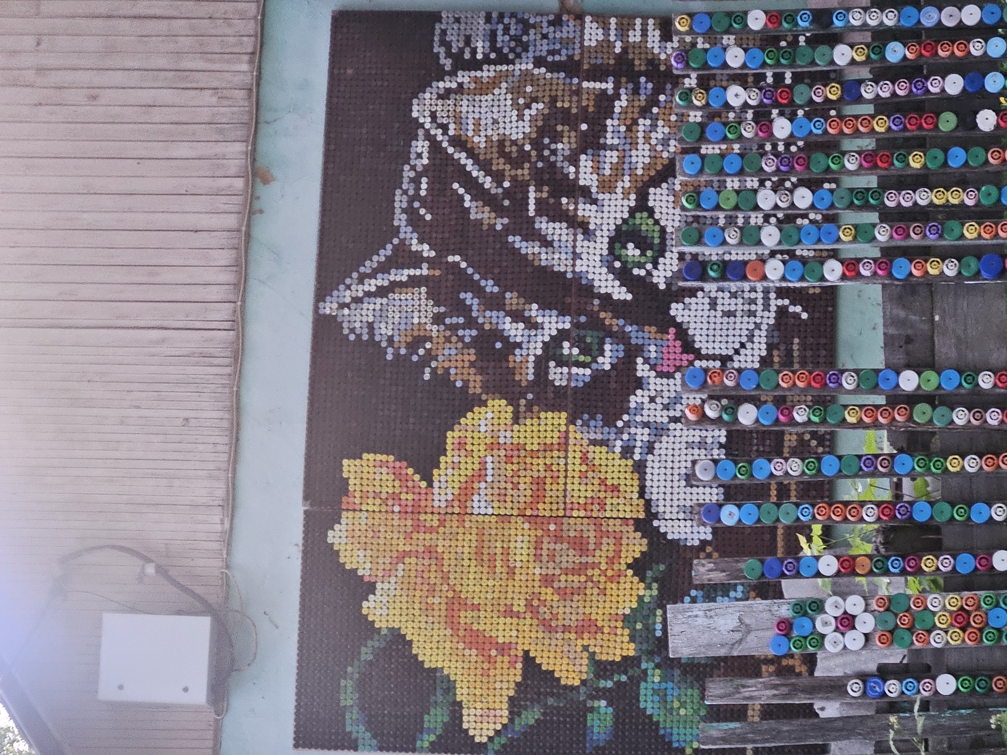 A mosaic artwork featuring a cat and a flower is visible on a structure, alongside horizontal rows of bottle caps mounted on wooden slats. The location is Rodniki, Russia. A white rectangular box is affixed to a wooden structure to the left of the mosaic. The artwork and bottle caps are positioned outdoors, with partial view of plant growth in the background.