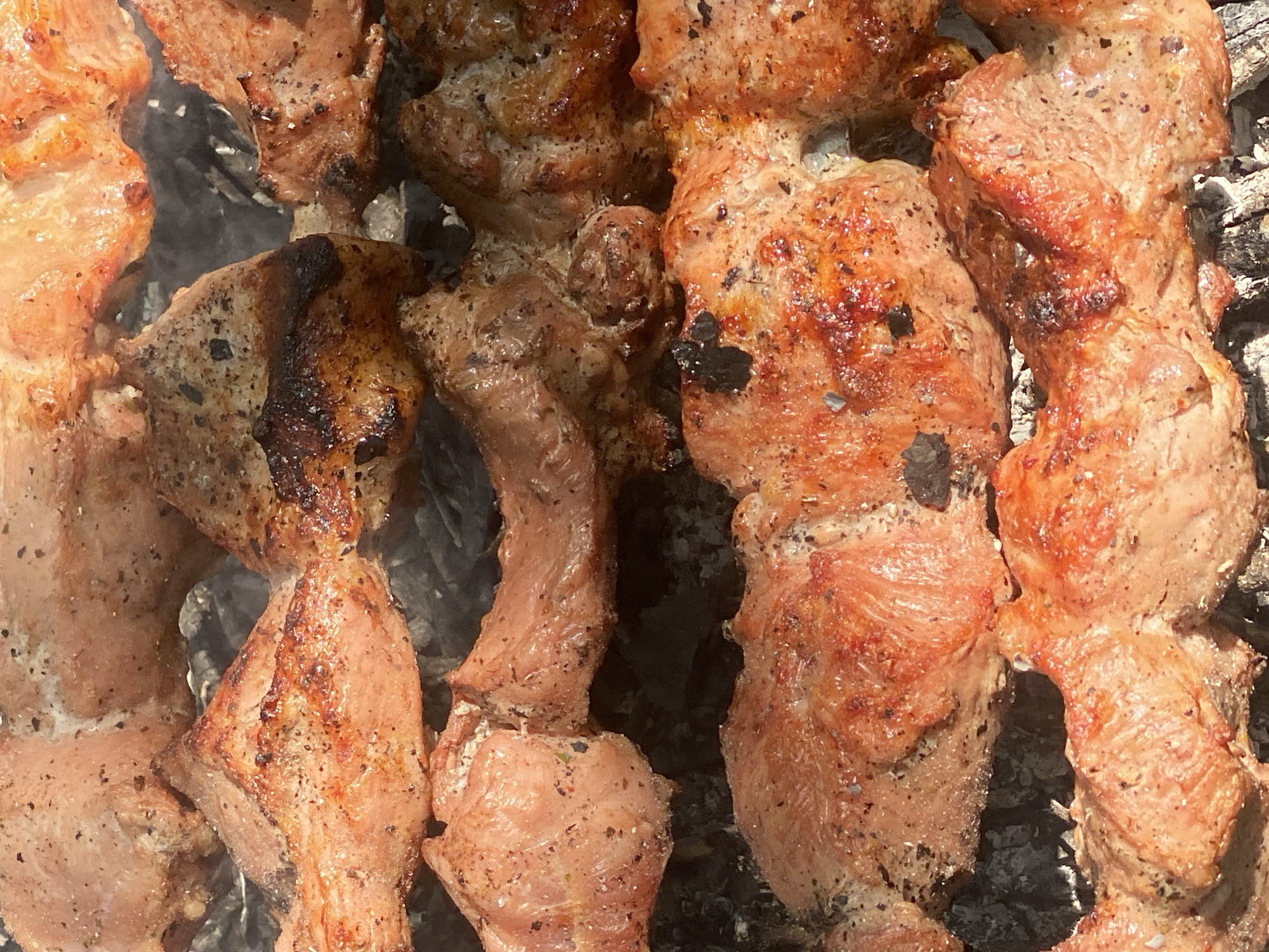 The image shows a close-up, overhead view of several skewers of seasoned meat, likely pork or beef, being grilled over charcoal. The meat is cut into bite-sized pieces and appears to be seared and slightly charred in places, indicating it is actively cooking. The background consists of glowing charcoal and ash. The lighting suggests an outdoor setting, possibly during daylight hours. There are no people visible in the image. The scene is consistent with a barbecue or outdoor cooking event. There is no information available to determine the specific location within Novokuznetsk, Russia.