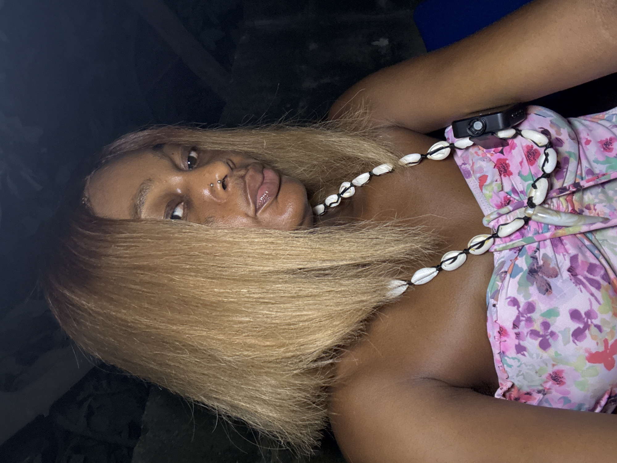 A medium close-up shot, angled diagonally, features a young woman lying down and looking towards the camera. She appears to be of African descent, with dark skin and long, straight, light blonde or golden-brown hair that spreads around her head. Her expression is direct, with a slight pout to her glossy lips. She has a double nose piercing, one a stud and the other a small hoop.
She is wearing a strapless or off-the-shoulder top, possibly a bikini top or a summer dress, adorned with a vibrant floral pattern in shades of pink, purple, green, and white. Around her neck and chest area, she wears a distinctive necklace made of cowrie shells, strung together with dark cord. A small, black electronic device, resembling a portable camera or recorder, is clipped securely to the upper part of her floral top near her right shoulder.
The background is dark and indistinct, suggesting a dimly lit indoor space or nighttime setting. A faint hint of dark blue is visible in the upper right corner, possibly a piece of furniture or clothing. The lighting on her face and upper body is soft, highlighting her features and the textures of her hair and accessories. No discernible text is visible in the image. The overall mood is relaxed and self-assured, consistent with a casual portrait. The location context is Ado, Nigeria.