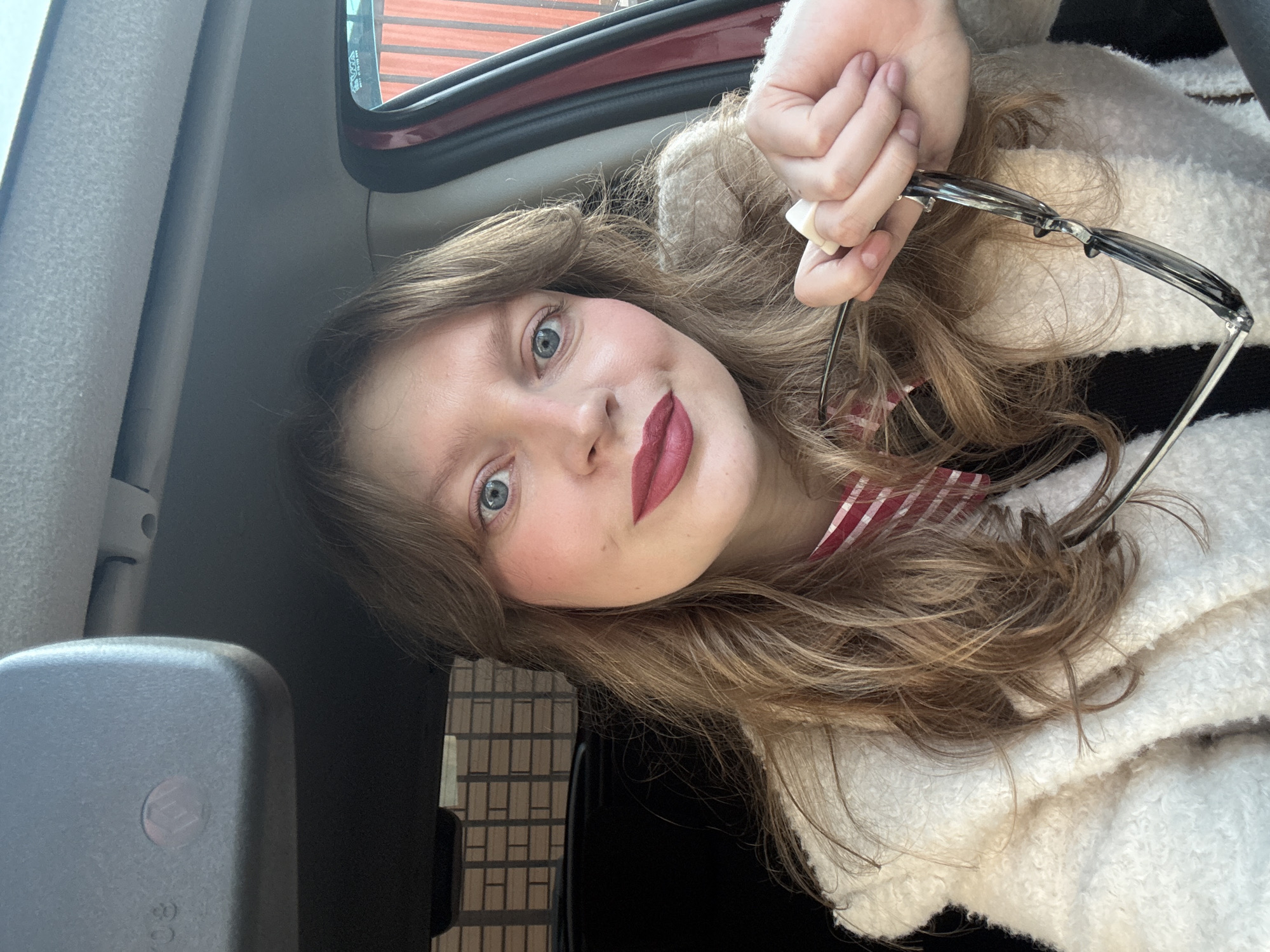 The scene captures a single female individual inside a vehicle in the city of Izhevsk, Russia. The subject, a fair-skinned woman with long, wavy light brown hair and blue eyes, directly faces the camera. She wears dark red lipstick and visible pink blush. Her attire consists of a cream-colored, textured, possibly fleece or boucle, outer garment over a red and white striped top. Her right hand, adorned with a white ring on a finger, holds a pair of dark-framed glasses. The vehicle's interior components visible include a light grey headliner, a grey rearview mirror with a subtle pinkish indicator light at the top-left, and a reddish-brown door frame on the right. Through the vehicle's windows, exterior elements are discernible: a light brown brick wall through the left rear window, and blurry, vertically oriented reddish-orange structures through the right window.