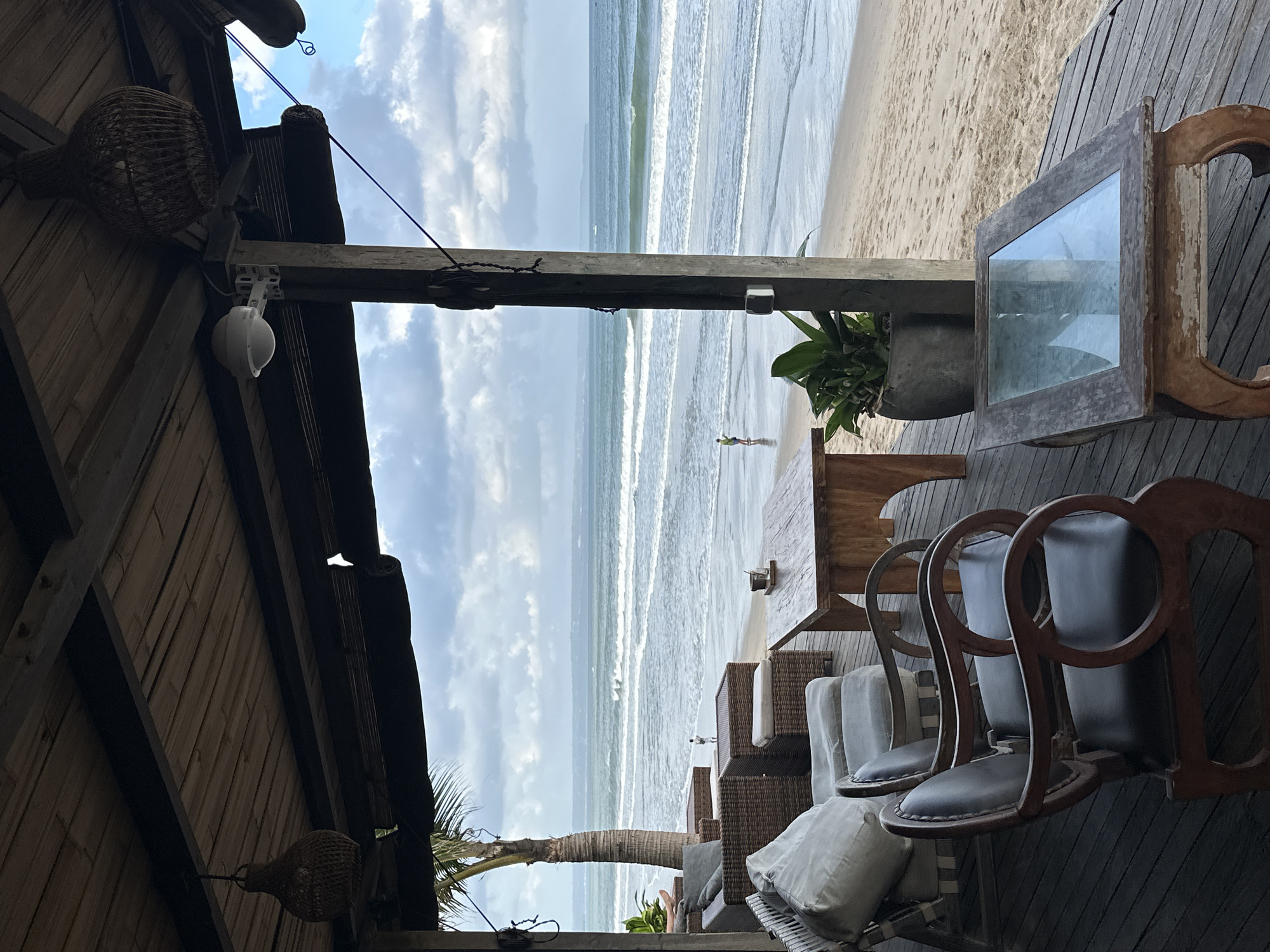 This image depicts an outdoor seating area at a beachfront establishment in North Kuta, Indonesia. The scene is viewed from a wooden deck under a thatched roof supported by wooden beams and pillars. In the foreground, two ornate wooden armchairs with dark leather seats are positioned on the deck, facing towards the ocean. To the left, a wicker sectional sofa with light-colored cushions is visible. A wooden coffee table with a glass top rests on the deck in front of the sectional. Further back, another wooden table with chairs and a planter containing a large green plant are present.
Beyond the seating area, the sandy beach stretches to the ocean, where gentle waves are breaking. A lone figure, appearing to be a person in a yellow shirt and shorts, is standing on the wet sand near the water's edge. The horizon shows a hazy coastline under a cloudy sky with patches of blue. Woven pendant lights hang from the thatched roof, and a security camera is mounted on a wooden pillar. The overall atmosphere suggests a relaxed, tropical setting suitable for dining or lounging with a view of the sea.