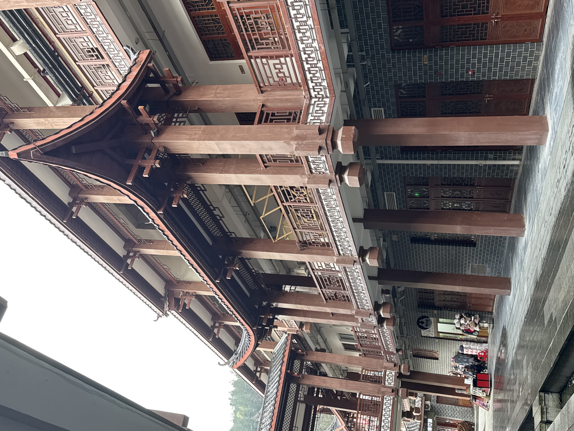 The image presents an outdoor, eye-level view of a traditional Chinese architectural complex in Wulingyuan, China, under an overcast sky. The scene features multi-story buildings constructed with dark grey brick facades on the ground floor and lighter walls above, adorned with extensive reddish-brown wooden elements. These include prominent square support pillars, intricate latticework railings on upper-level balconies and walkways, and characteristic curved, tiled roofs.
The pavement in the foreground is wet and reflective, indicating recent rain. On the ground level, several closed dark wooden doors with decorative latticework are visible. One storefront on the left is open, displaying merchandise outdoors: various garments on racks, a red display cart, a mannequin head adorned with hats, and a rack holding stuffed animals. A circular dark sign is mounted above this shop's entrance. Numerical labels "330" are visible on an upper floor doorframe, and "230" on a ground floor door. Green, forested hills are discernible in the distant background on the left side, above the rooftops. A narrow, concrete-lined drainage channel containing some debris is present in the immediate lower-left foreground. No individuals are present within the frame.