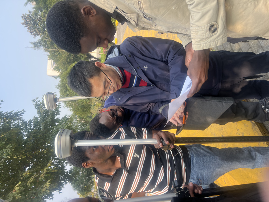 Four people are clustered together, seemingly focused on paperwork held by the person in the center. Two individuals are holding devices mounted on poles, possibly surveying equipment. The clothing styles suggest diverse backgrounds. A tree and a building are faintly visible in the background. The scene is bathed in daylight, creating a slightly hazy atmosphere. A yellow painted area lies on the ground.