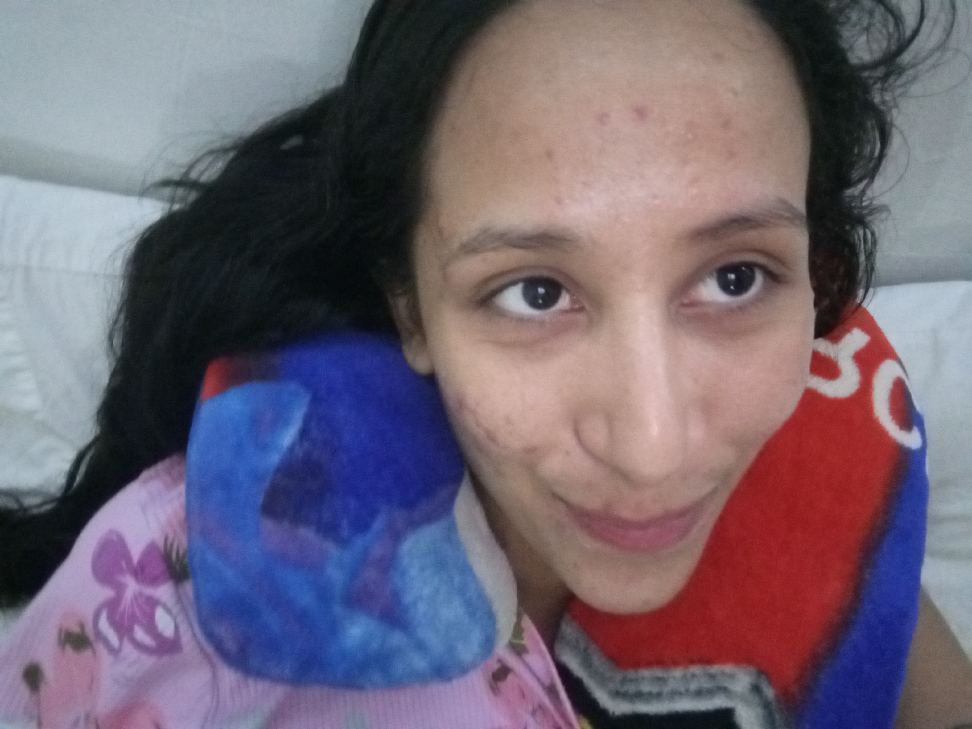 A woman is lying in bed with her head resting on a red, white, and blue pillow. She has long, dark hair and is looking to the right of the frame with a slight smile. She is wearing a pink and white shirt with a floral pattern. A blue pillow is on her left. The background is a white pillow or sheet. The location in the city of Porlamar, Venezuela is not discernible from the image.
