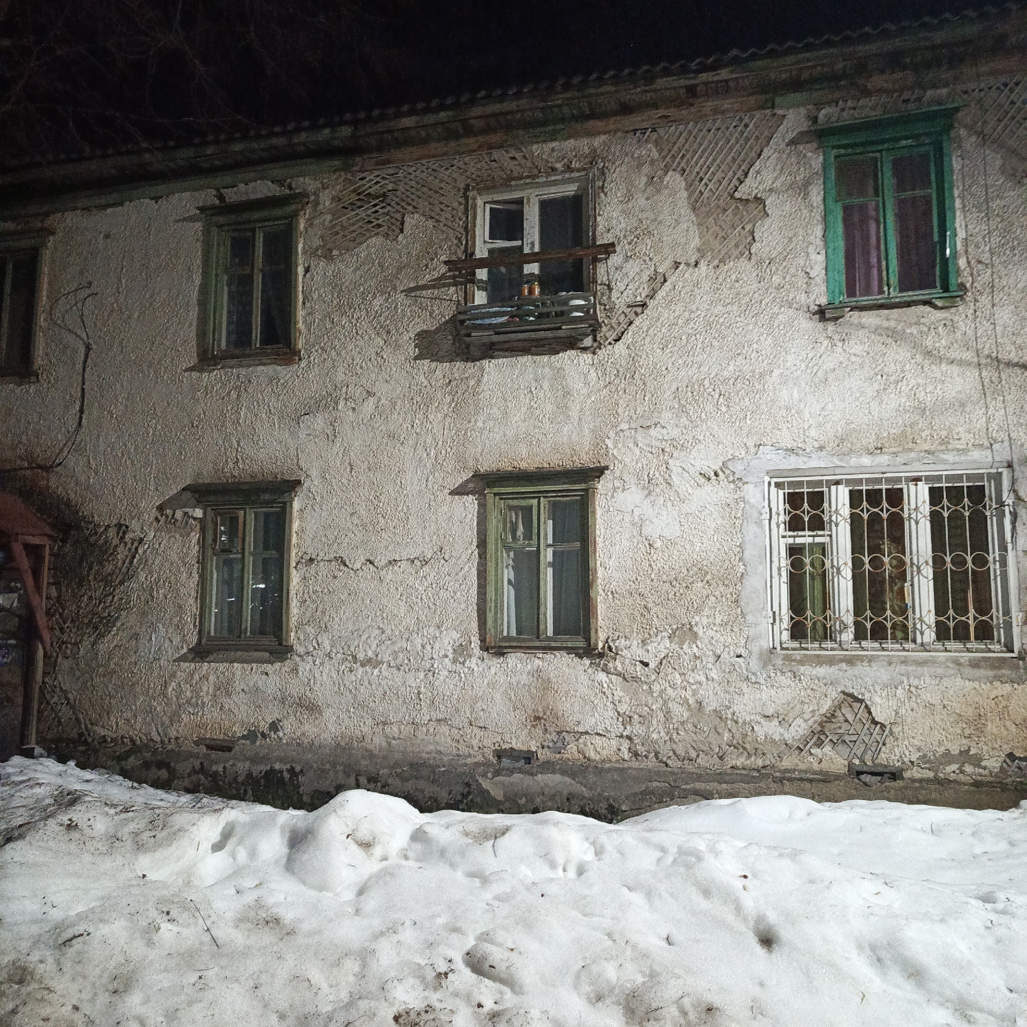 The image depicts the facade of an old, two-story building in the city of Chaikovsky, Russia. The building has a rough, textured off-white exterior. The windows are framed in various shades of green, except for one which has a white frame and bars. A small, makeshift balcony protrudes from one of the upper windows. In front of the building, there is a large mound of snow, suggesting a winter setting. The scene is illuminated at night, possibly by streetlights. A partial wooden structure is visible to the left, possibly a porch or shed.