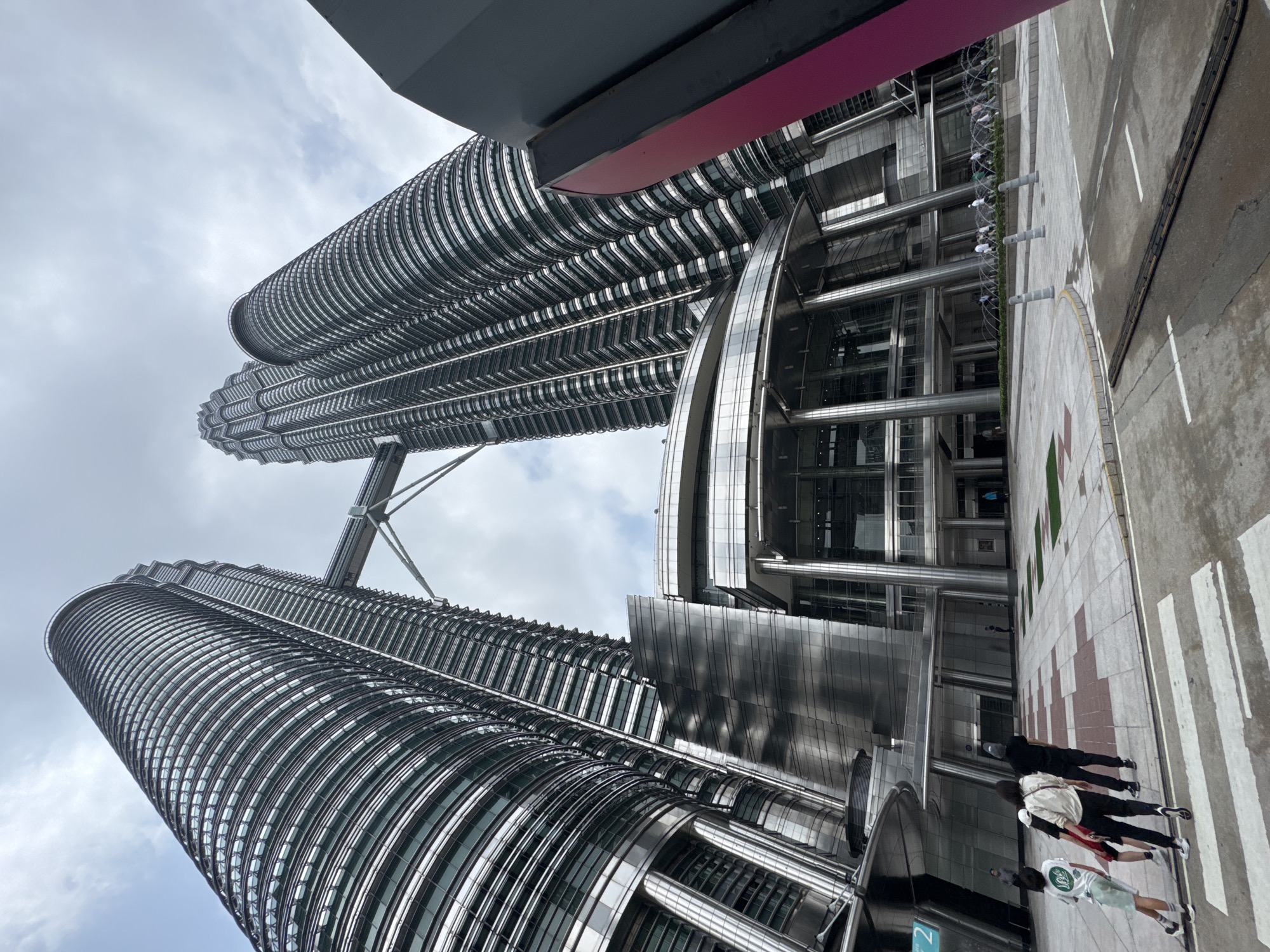 The image shows the Petronas Twin Towers in Kuala Lumpur, Malaysia, from a low-angle perspective. The towers are constructed of metal and glass, with a distinctive cylindrical design. A skybridge connects the two towers at a high level. In the foreground, several people are walking along a sidewalk. The sky is overcast. A partial view of a building or structure with a magenta accent is visible above the towers. The street and sidewalk are clean and well-maintained.