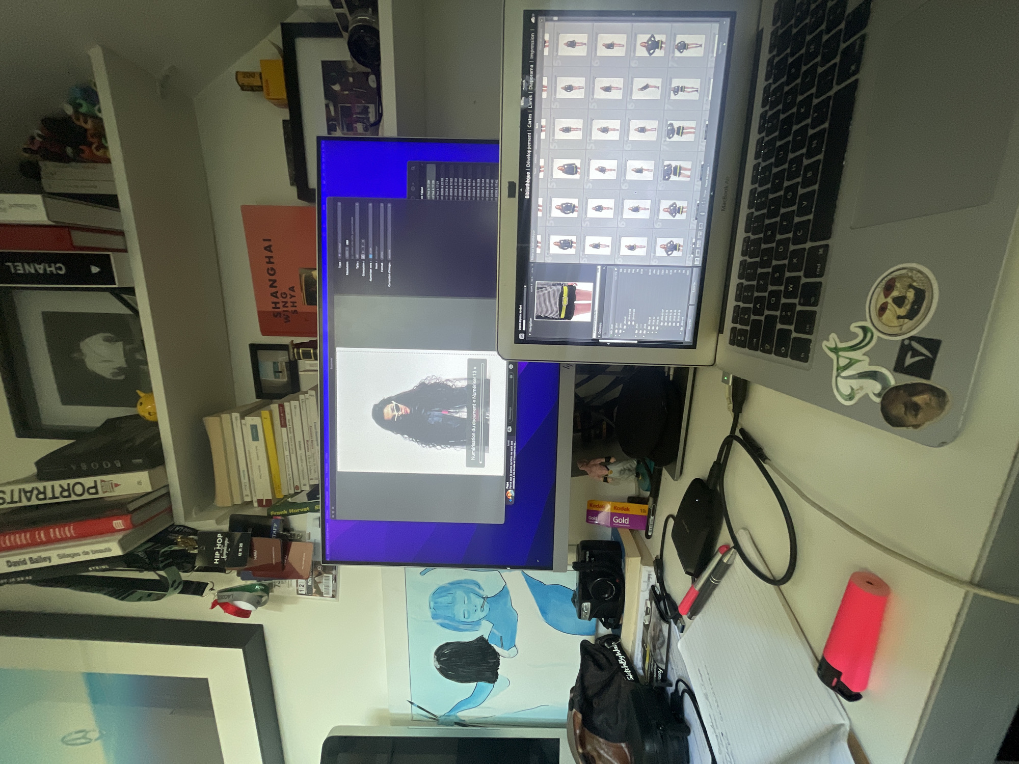 The image displays an active, well-lit workspace, likely a home office or creative studio, filled with computing equipment, books, and various personal items. The primary subjects are two computer screens—a MacBook Air laptop and an external monitor—positioned side-by-side, suggesting a multi-monitor setup for productivity.
On the external monitor, an application interface is visible, showing a large image of a woman with long, dark, curly hair and sunglasses. Accompanying text, predominantly in French, includes "Numérisation du document « Numériser 13»" (Document digitization « Scan 13»), indicating an image processing or scanning activity is underway. The laptop screen displays a photo management software (resembling Adobe Lightroom) with a grid of numerous thumbnail images of models, with one larger selected image of a person in a striped top and yellow belt. Interface elements like "Bibliothèque," "Développement," "Cartes," "Livres," "Diaporama," and "Impression" are visible, all in French.
The desk surface around the monitors holds an assortment of objects, including an external hard drive, charging cables, a pink highlighter, a pen, a small notebook, a camera (possibly a film camera given the Kodak Gold film rolls nearby), and a small painting featuring two stylized blue figures. The MacBook Air has several stickers, including one with a skull design and another abstract green emblem.
To the left and behind the desk, several bookshelves are packed with books. Visible titles include "CHANEL," "SHANGHAI WING SHYA," "PORTRAITS," "BOOBA," "David Bailey Silages de beauté," "HIP HOP," and "Frank Horvat." A small yellow toy is also perched on a shelf. The scene is indoors, well-lit, suggesting daytime or bright artificial lighting. The overall impression is a vibrant, creative environment where digital and physical media coexist. The text visible on the screens and books confirms the French context, aligning with the provided location context of Paris, France.
