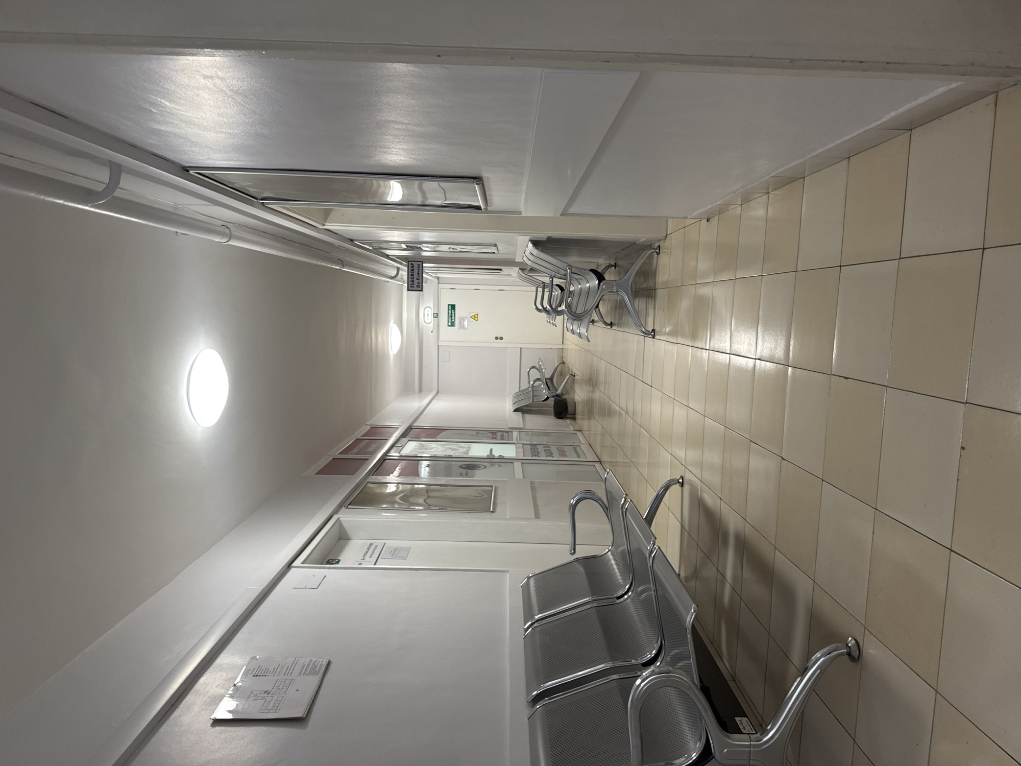 The image depicts an interior corridor, likely a waiting area in a public building, possibly a hospital or clinic. The location is in Plovdiv, Bulgaria. The corridor has light-colored tiled flooring and white walls. Rows of metal benches line the corridor on both sides. There are several doors along the left wall, with informational signs and posters. Overhead, there are fluorescent lighting fixtures and various pipes or conduits. The perspective is from one end of the corridor, looking towards the other, and the scene is empty of people. A fire extinguisher is placed by the exit, indicated by the international sign.