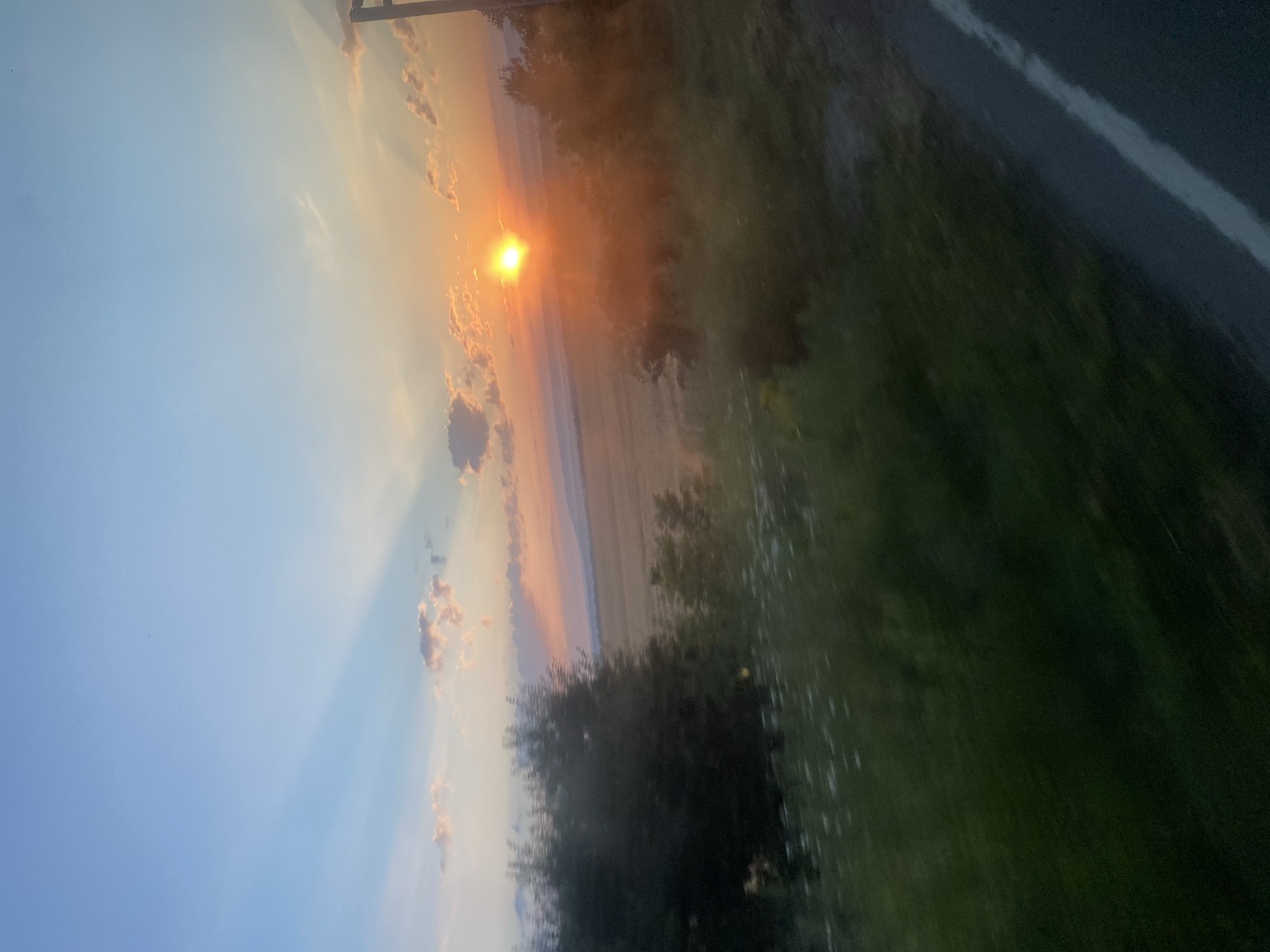 The image depicts a landscape scene at sunset. The sun is visible as a bright orange orb low on the horizon, casting rays through the sky. The sky transitions from a clear blue at the top to hues of orange and pink near the horizon, with scattered clouds. In the foreground, there is blurred greenery and a road with a white line marking. The background shows rolling hills and fields, possibly farmland, with some trees visible. The overall impression is of a rural setting during golden hour, with the motion blur suggesting the photo was taken from a moving vehicle. This location is identified as Novosiolovca, Moldova.