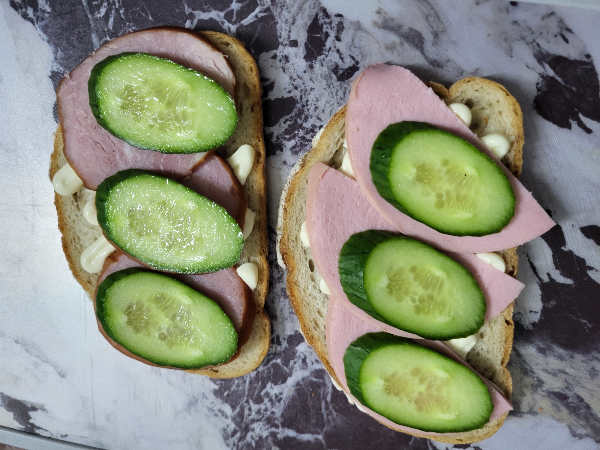 The image displays two open-faced sandwiches on a marble-patterned surface. Each sandwich consists of a slice of bread topped with mayonnaise, slices of ham or a similar deli meat, and several slices of cucumber. The sandwiches are arranged vertically, one above the other, with the top sandwich positioned slightly to the left. There are no people or other discernible objects in the scene. The location of Horishni Plavni, Ukraine is not depicted or suggested by any elements within the image.