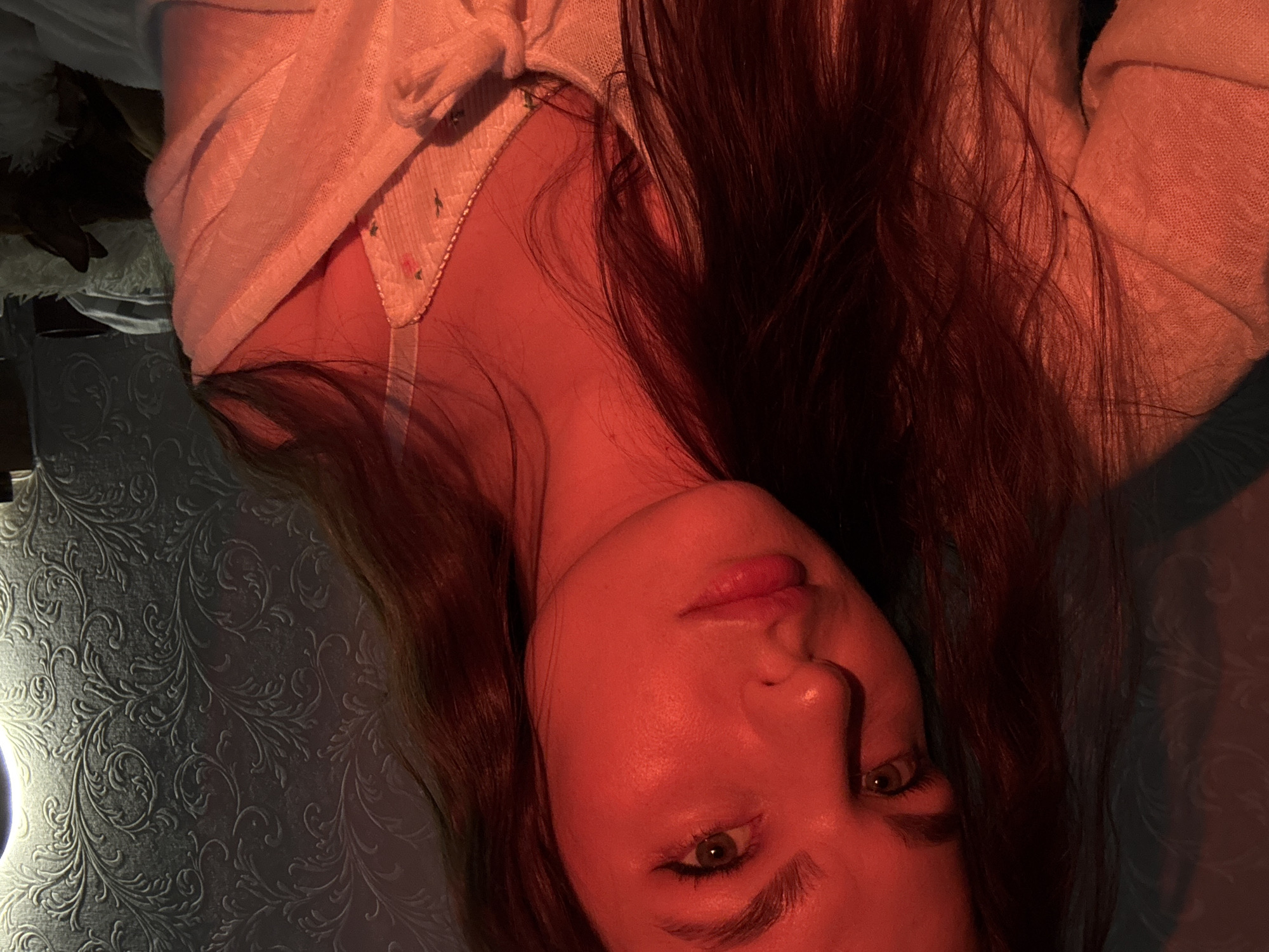 A woman with long, brown hair and green eyes is laying on a patterned blue surface. She is wearing a white top with a visible white bra underneath. The scene is lit with a warm, reddish light. The location in Chaikovsky, Russia, is not discernible from the image.