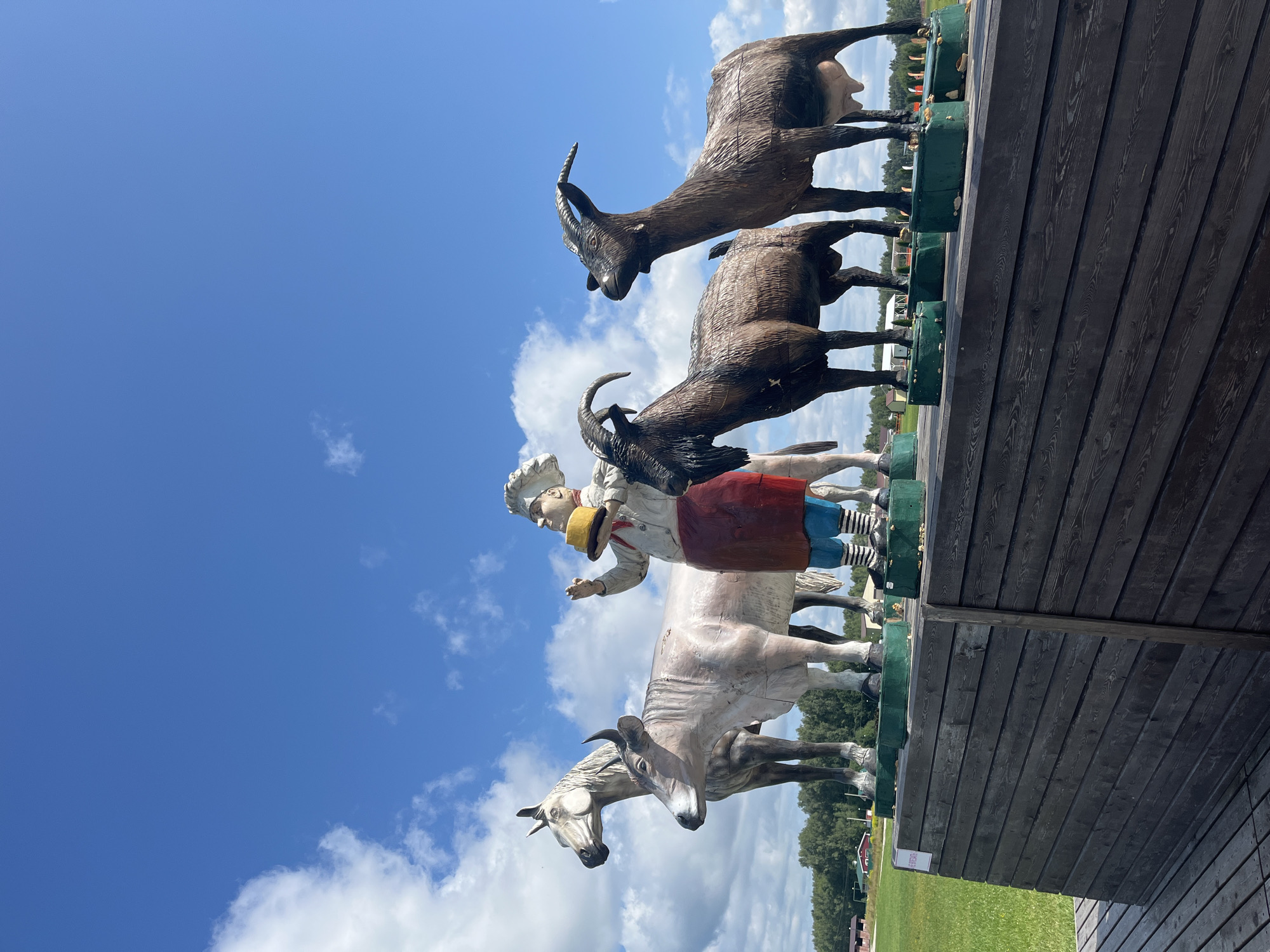 This is a full outdoor shot of several large, sculpted figures displayed on a wooden platform. The figures, positioned in a line from left to right, include a pale horse, a cow, a chef holding a round yellow object, a dark brown goat with horns, and another dark brown goat with horns and visible udder. The sculptures appear to be made of a textured material and are brightly colored. The background features a bright blue sky with scattered white clouds, a grassy field, and a line of trees with some buildings in the distance. A portion of a dark wooden deck is visible in the foreground, leading up to the platform where the sculptures are displayed. The scene is located in the village of Akhlebaikha (Kovriginskii S/S), Russia.