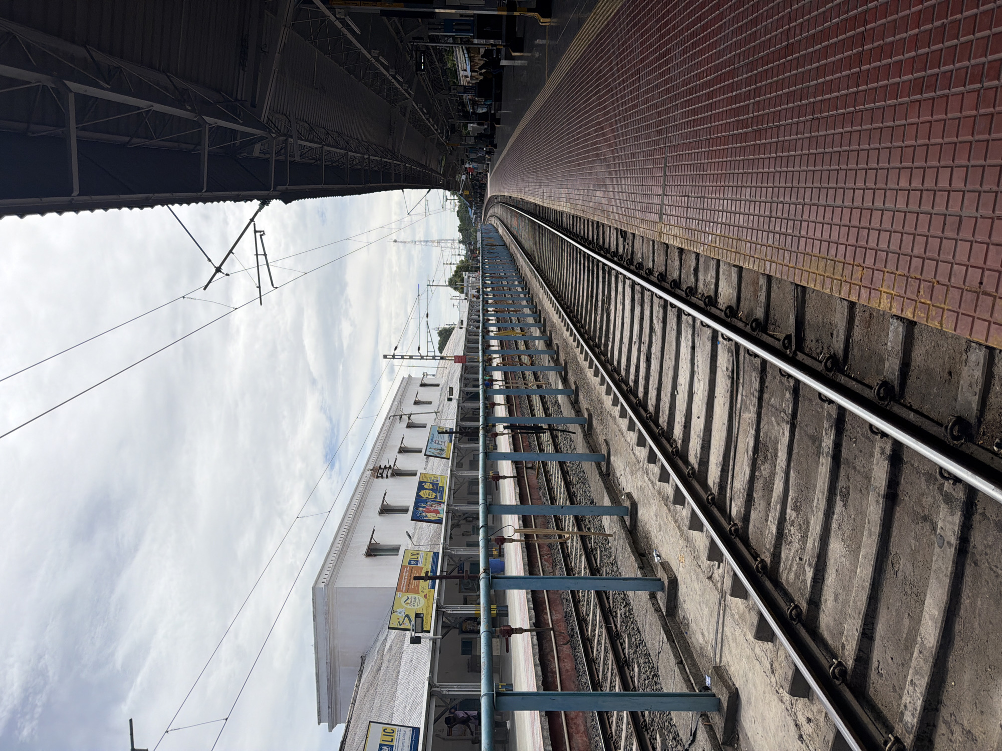 This image captures a railway station platform in Thanjavur, India, with a distinct upward perspective. The scene is dominated by the curved railway tracks leading into the distance, bordered by a red tiled platform on the right and a covered walkway with metal support structures on the left. Overhead wires are visible against a cloudy sky.
A long, white building with multiple windows lines the platform. Several billboards are affixed to its exterior, featuring advertisements and information, some with legible text. One billboard prominently displays "LIC" in yellow and black. Another appears to advertise insurance or financial services with images of people. The architecture of the building is simple and functional, typical of many Indian railway stations.
The time of day appears to be daytime, with diffused sunlight filtering through the clouds. The weather seems overcast. The presence of the railway tracks, platform, and station building clearly indicates the setting is a train station. A few individuals are visible on the platform near the building, but they are small in the frame and not the primary focus. The overall mood of the image is neutral, conveying a sense of everyday transit and the infrastructure supporting it. The perspective emphasizes the expanse of the railway lines and the architectural elements of the station.