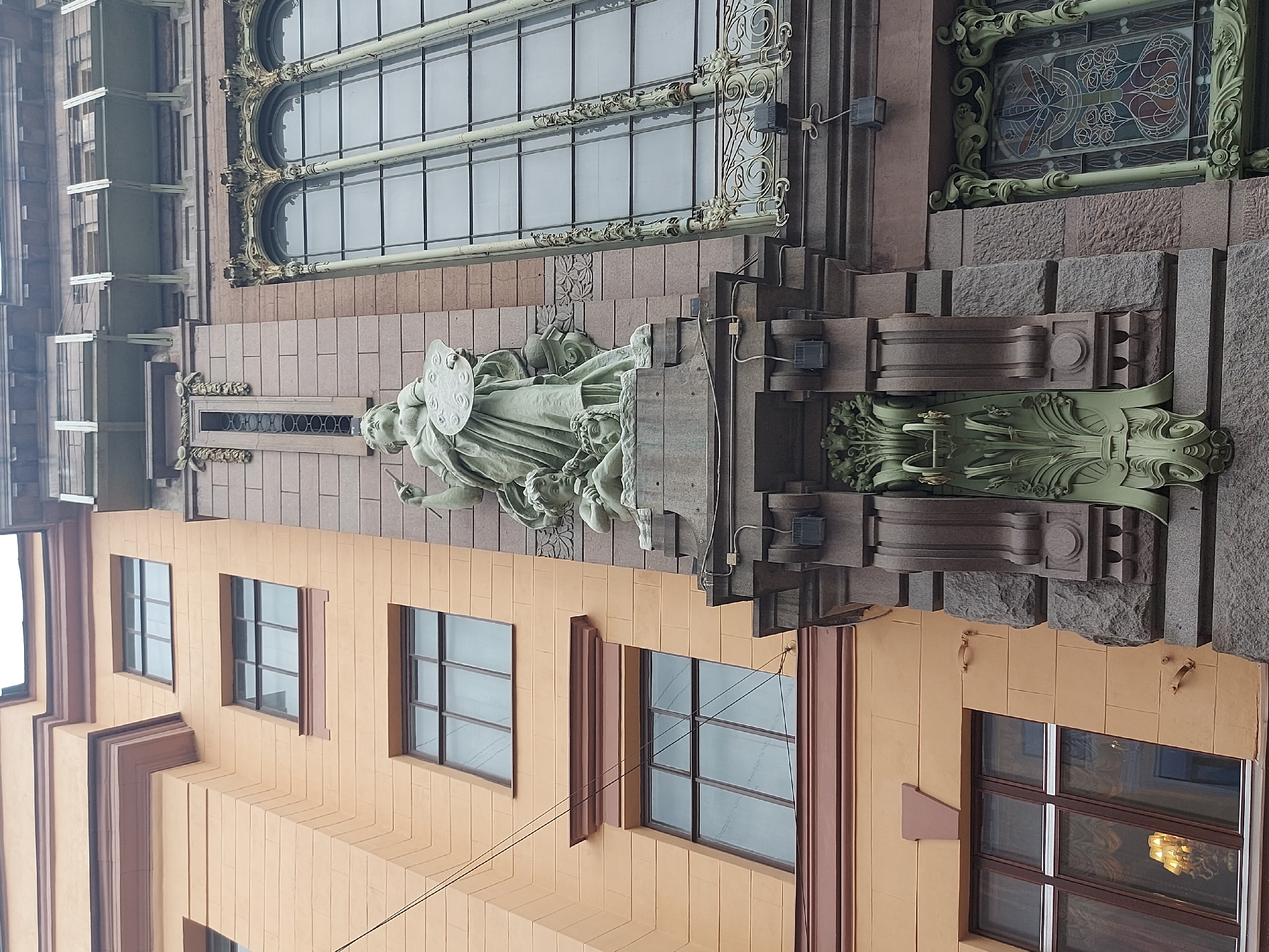 The image shows a building facade in Saint Petersburg, Russia, specifically the facade of the Singer House (also known as the House of Books). The building is characterized by its Art Nouveau style.
Visible architectural elements include:
* Large windows with ornate metal framing
* A statue of Minerva (Roman goddess of wisdom and crafts) holding a palette
* Decorative green sculptural elements below the statue
* A multi-layered facade with sections of brown and tan-colored stonework
* Visible windows with frames of brown and tan colors
The scene is likely taken from street level, looking upwards at the building’s facade.