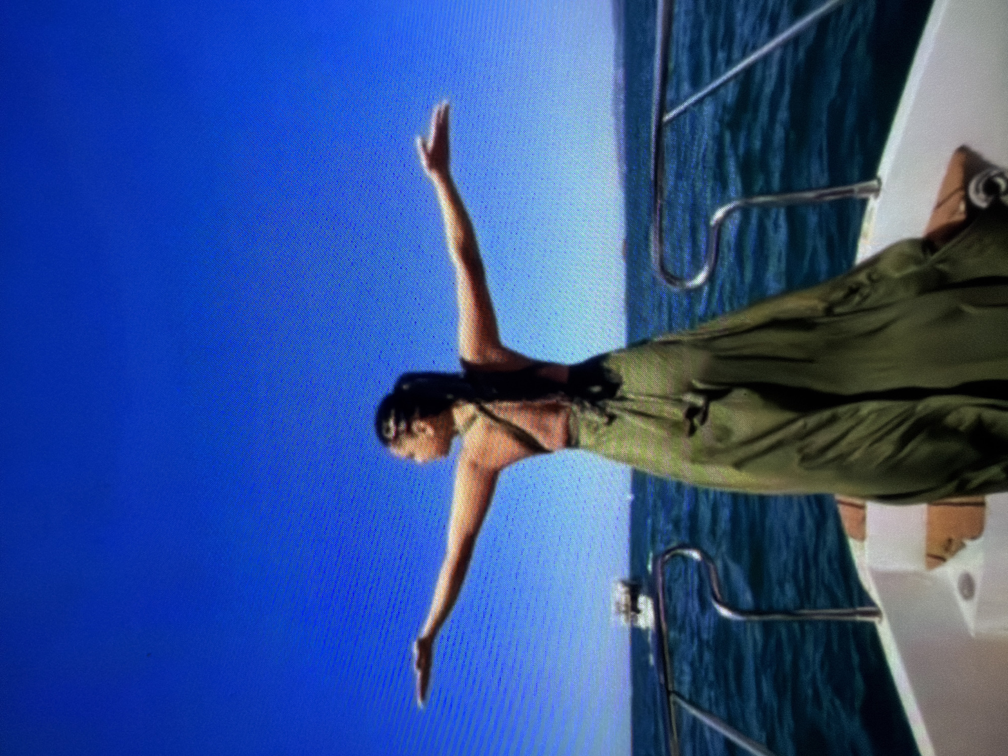 A woman in a long olive-green dress stands with arms outstretched on a white boat, presumably at sea as indicated by the blue water and clear blue sky in the background. A silver railing is visible on the boat. The location is unknown.
