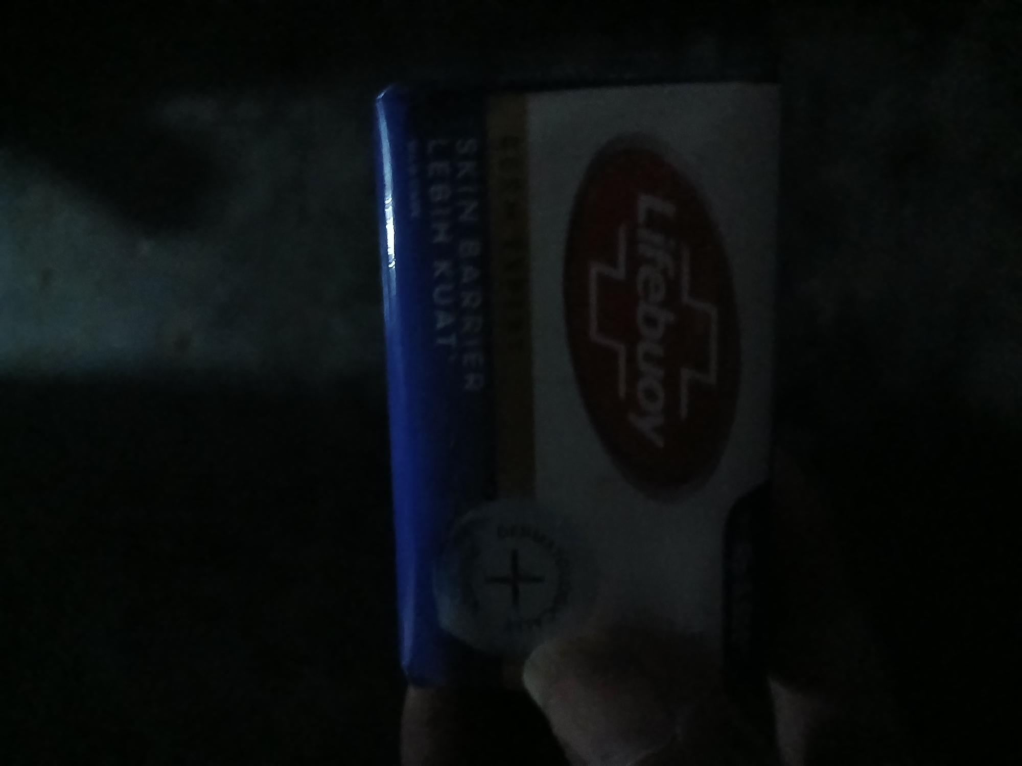 The image shows a close-up of a Lifebuoy soap bar packaging, held in a hand against a dark background. The packaging is blue and white, with the "Lifebuoy" logo visible in red within an oval. Text on the packaging is partially visible, including "SKIN BARRIER" and "LEBIH KUAT". The lighting is dim, and the background is predominantly dark and out of focus, suggesting an indoor environment. There is no discernible information about a specific location in the city of Kademangan Kulon, Indonesia.
