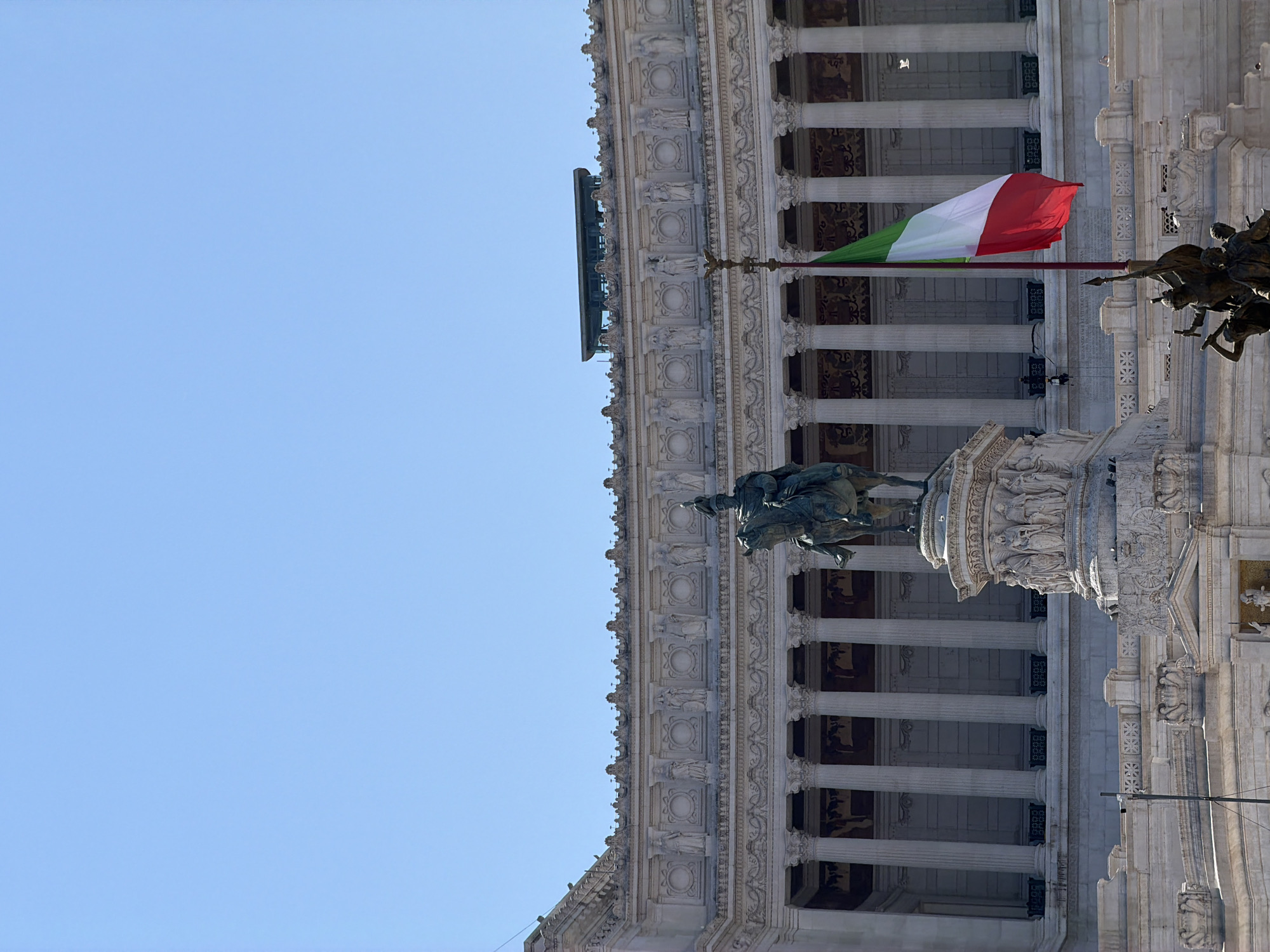 A wide shot captures a monument against a clear blue sky and a building. The monument features a bronze equestrian statue of a man in a military uniform. The statue stands on a tiered pedestal adorned with carved figures. To the right of the statue, an Italian flag flies from a tall pole. The background shows a large, ornate building with classical columns and decorative architectural elements. A few small figures can be seen on the building's facade and in front of it.