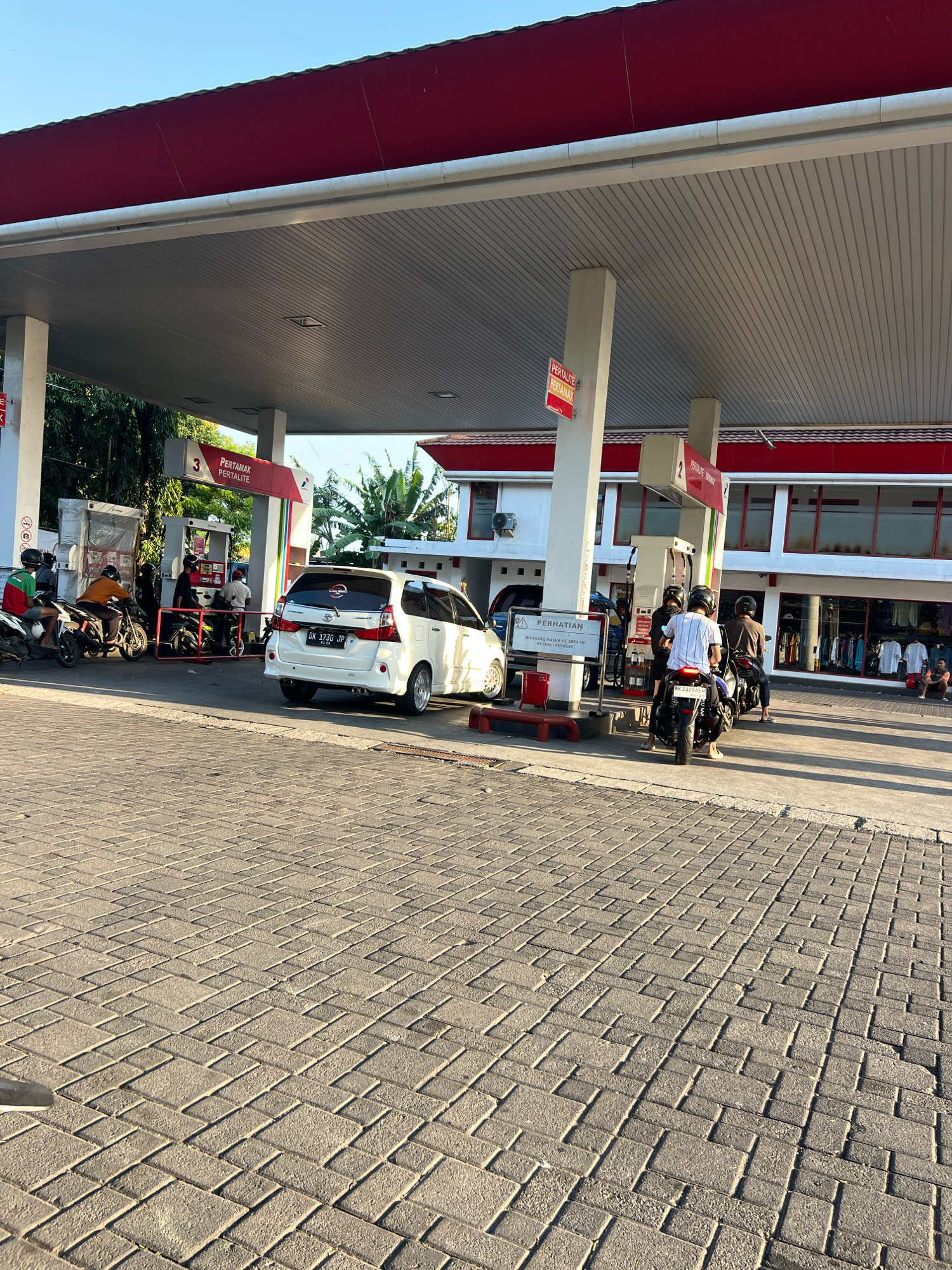 The image depicts a gas station in Denpasar, Indonesia, under a bright, clear sky. The gas station features a prominent red roof supported by white pillars. Several fuel pumps are visible, with a white minivan at pump number 3 and motorcycles at pump number 2. People are seen refueling their vehicles and interacting at the pumps. In the background, a building with red trim and large windows suggests a convenience store or service area. The ground is paved with light-colored interlocking bricks. Trees and foliage are present in the surrounding environment.