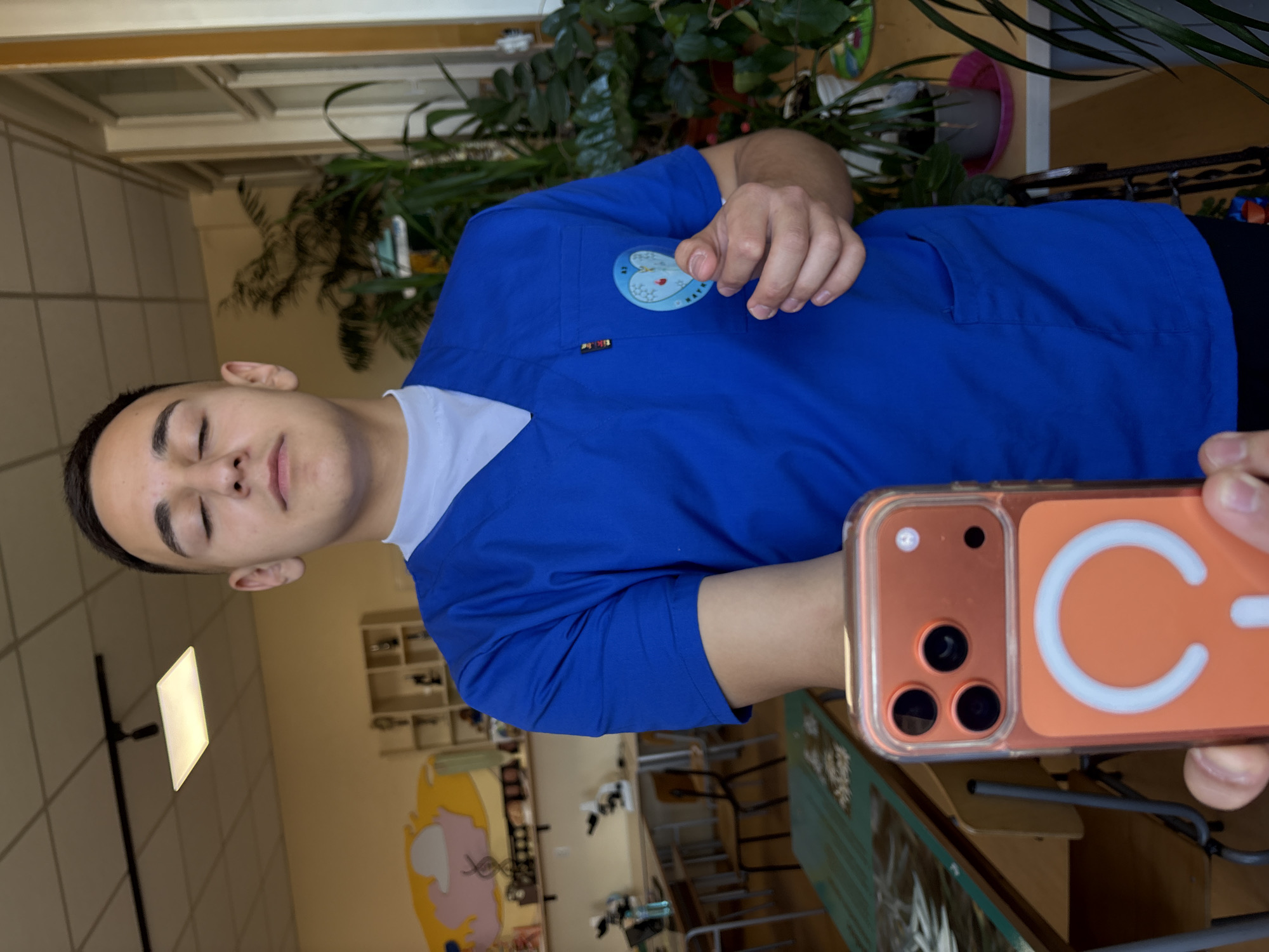 A young man in blue scrubs stands indoors, holding an orange phone with a white C-shaped logo up to take a selfie. The room contains a desk and shelves with objects. In the background, there is a wall with a mural. The location appears to be in Panagyurishte, Bulgaria, based on a patch on the scrubs indicating "Panagyurishte." There are plants in the background as well.