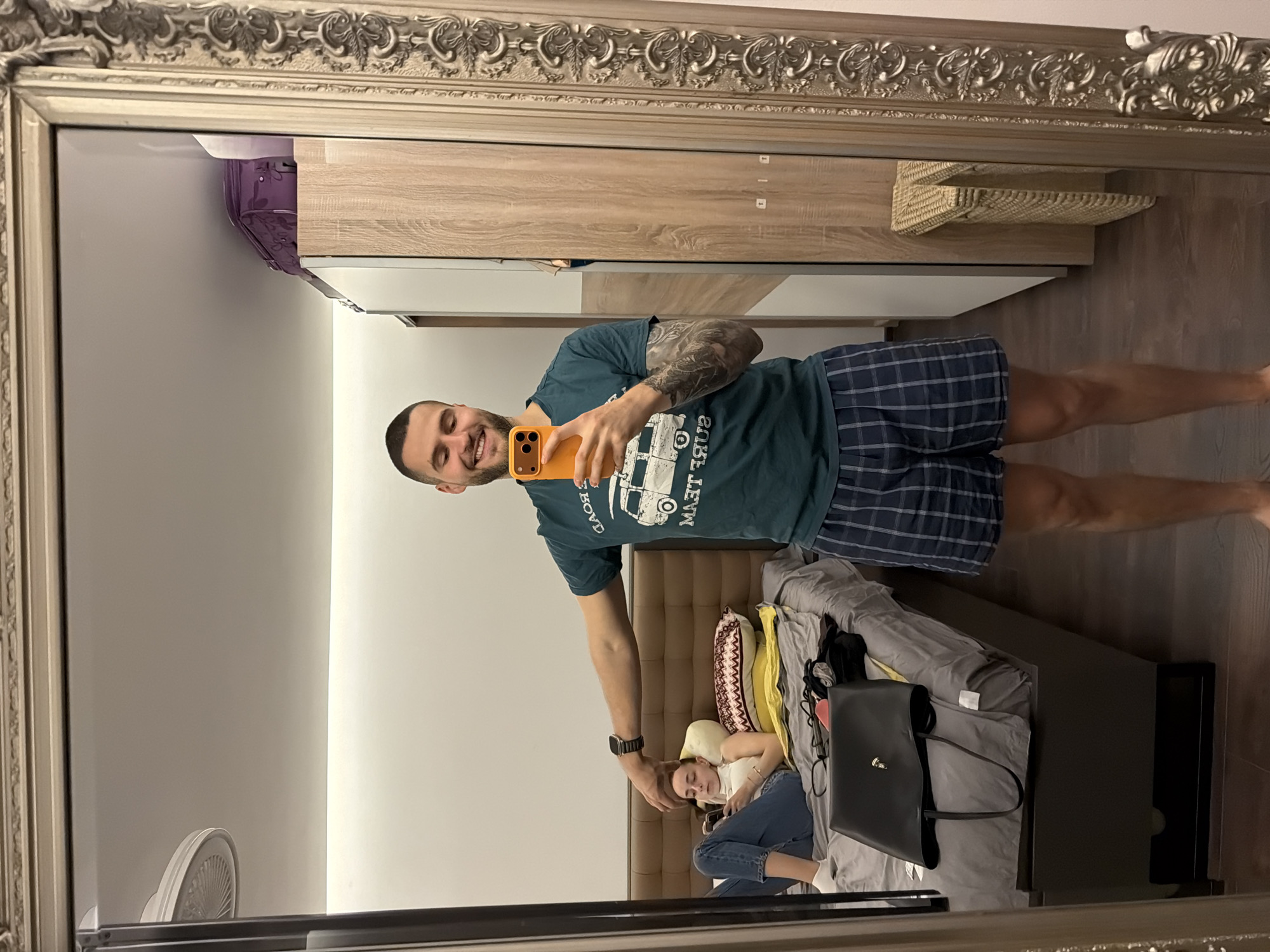 This mirror selfie, taken in an indoor setting in Sofia, Bulgaria, captures a man and a woman in a casual, relaxed moment. The image is reflected in an ornate silver-framed mirror, showcasing a room with wooden flooring and light-colored walls.
The primary subject is a man positioned centrally, taking the photo with an orange-cased smartphone. He has short dark hair, a beard, and distinctive tattoos on his left arm. He is wearing a dark teal t-shirt featuring a white graphic of a camper van and the mirrored text "GLOBO TRAVEL". He also wears blue plaid shorts and a watch on his left wrist. The man is smiling broadly, conveying a sense of happiness or contentment.
In the background, a woman is relaxing on a bed with a brown padded headboard. She has light-colored hair and is dressed in a white top and blue jeans, appearing to be looking at a light-colored smartphone or resting. The man's left hand is gently placed near her head, suggesting an affectionate gesture.
Other visible objects include a wooden wardrobe or cabinet with a purple container and a woven shelf on top, reflected in the upper right. A white circular fan is visible in the lower left of the reflection. A black handbag and a partially visible laptop are also situated on the bed near the woman. The scene depicts a comfortable, daytime atmosphere.