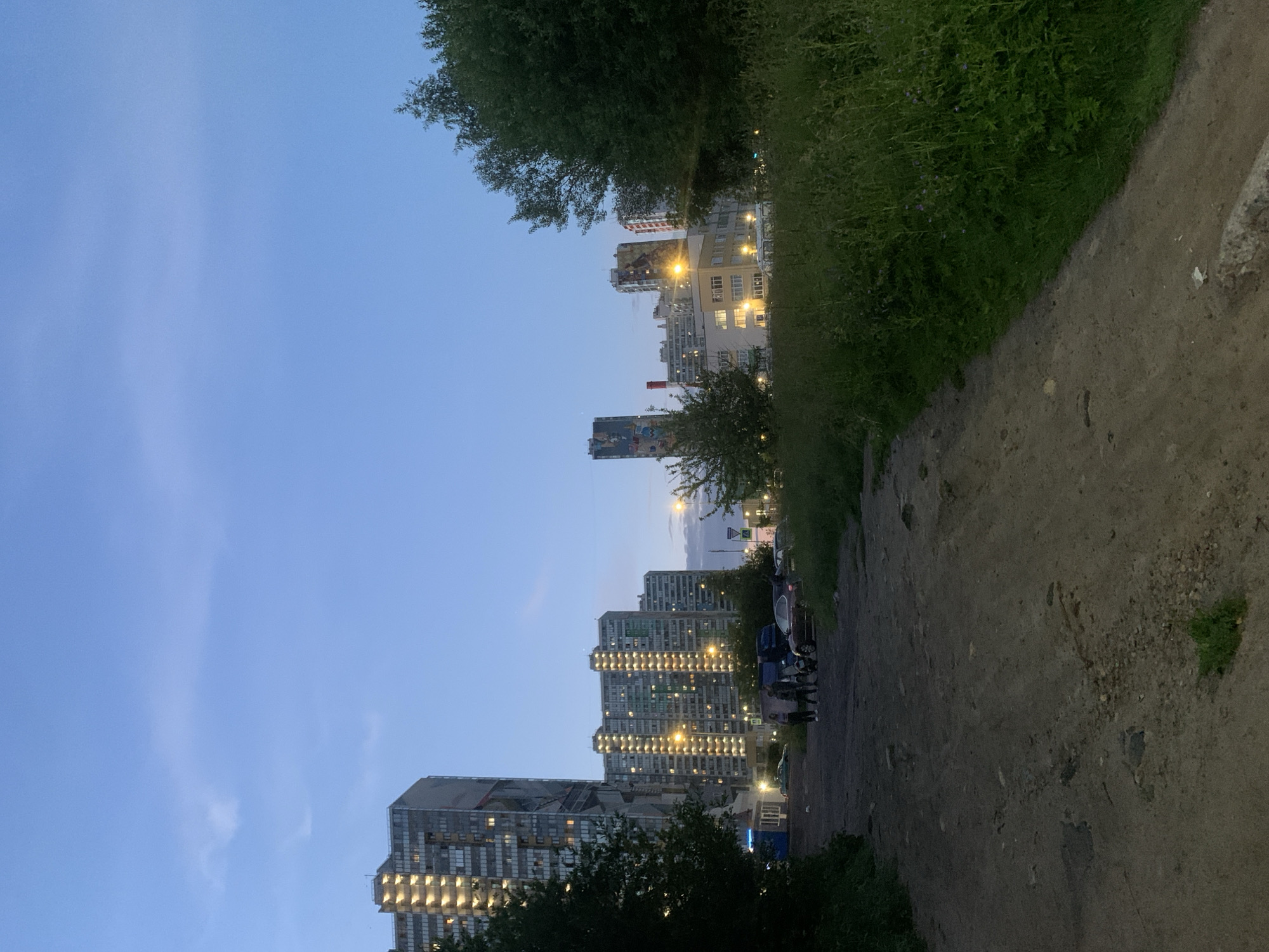 The image depicts a twilight scene in a residential area of Saint Petersburg, Russia. A dirt path leads towards several high-rise apartment buildings, some of which are illuminated with interior lights. The sky is a soft blue, transitioning from day to night, with faint clouds visible. Tall, modern buildings dominate the background, with the architecture suggesting a contemporary urban setting. In the mid-ground, a few individuals are standing near parked cars, suggesting casual gathering or departure. The foreground is characterized by the unpaved path and overgrown green vegetation, indicative of a less developed or more natural area within the city. The overall impression is one of an urban periphery at dusk.