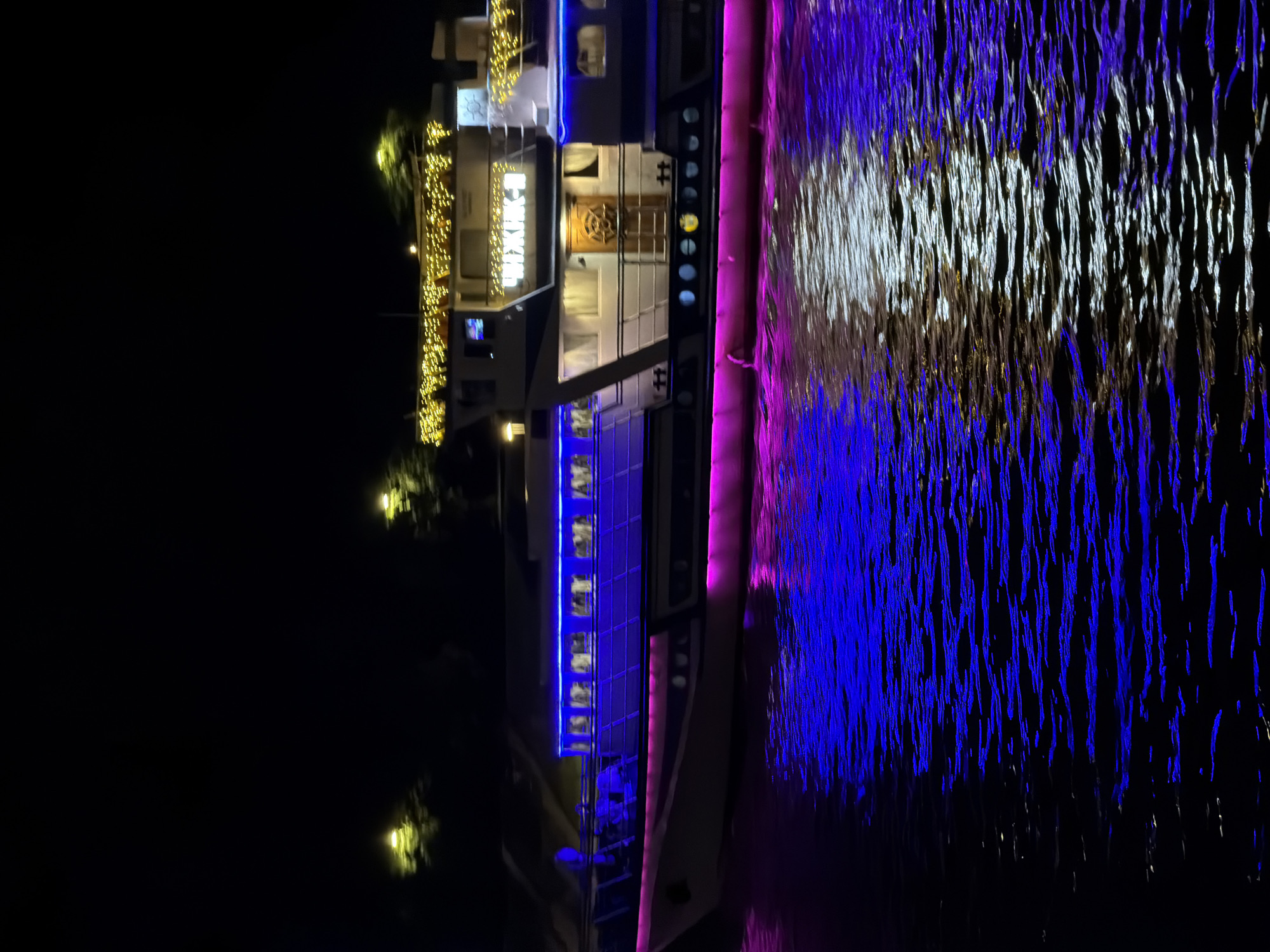 At night, a brightly illuminated riverboat or ship is docked on a body of water that reflects the lights. The lights on the boat include:
* Rows of golden string lights outlining the top deck and other areas of the boat.
* Blue LED lighting running vertically along the boat's structure, possibly on the windows or sides.
* A distinct and ornate round wooden door or decorative element is visible on the boat.
* The surface of the water reflects these lights in a shimmering, distorted manner. There are white and blue reflections indicating the different light sources from the boat.
It's not possible to pinpoint the specific location within Moscow or to determine if this is in Moscow solely from the image.
